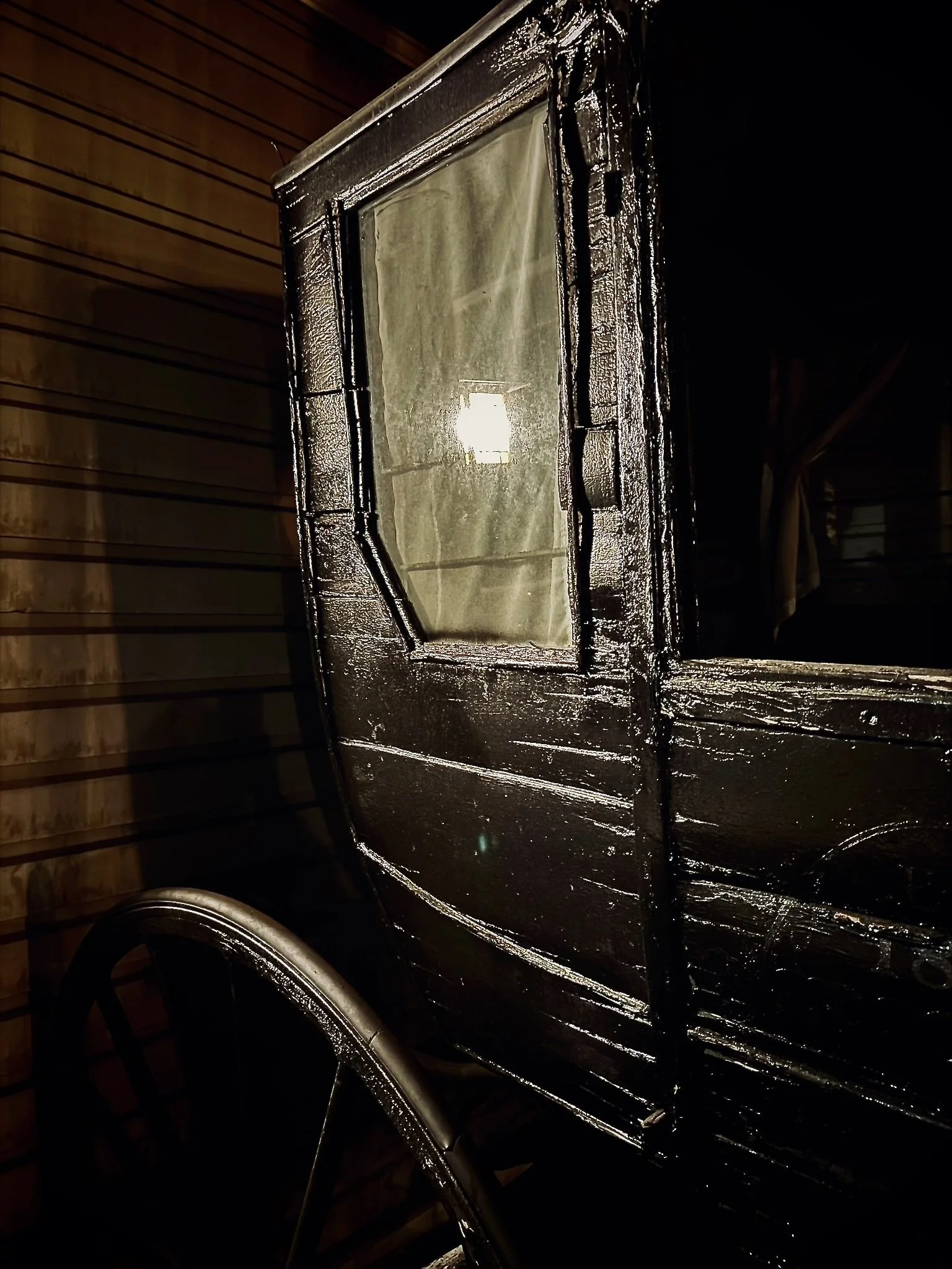 Close-up of a black vintage carriage with a window, reflecting a light source on its shiny surface, against a wooden wall background.