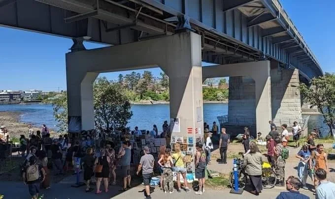 Crowd of people gathering under a bridge near a body of water, with some sitting on benches and others standing, at an outdoor art event.