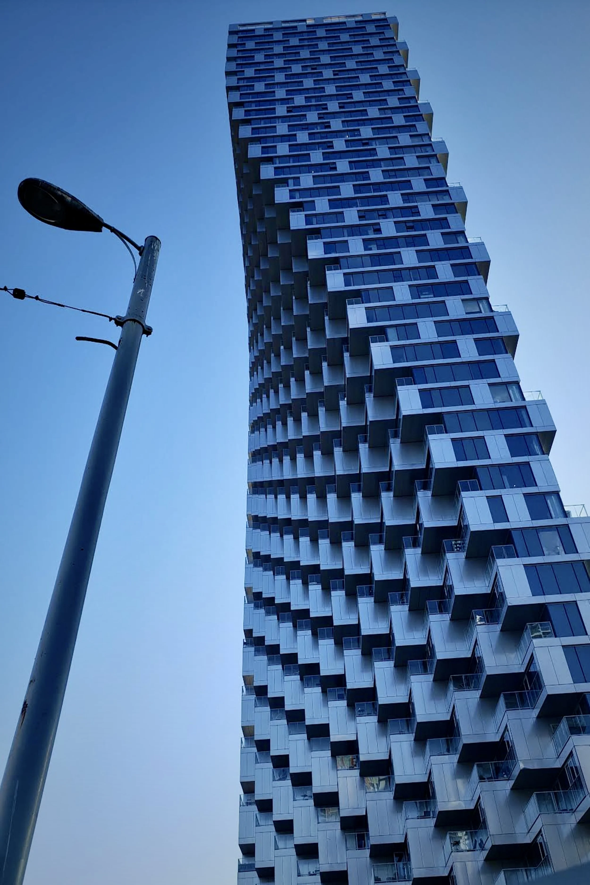A modern high-rise building with a unique, irregular facade and multiple balconies, photographed from a low angle against a clear blue sky, with a street lamp in the foreground.