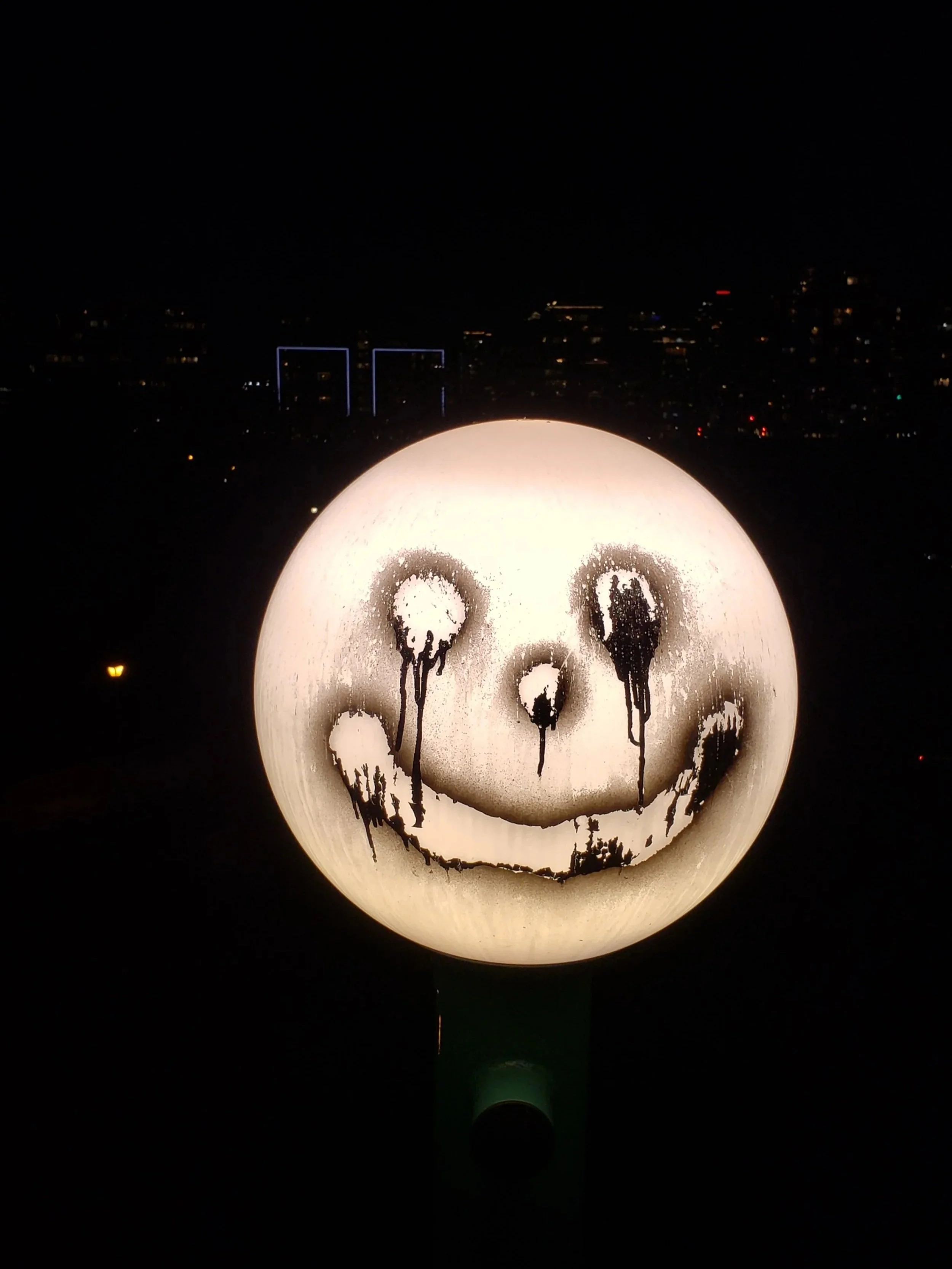 Spherical outdoor light with a smiley face drawn on it using black spray paint, placed against a nighttime cityscape background.