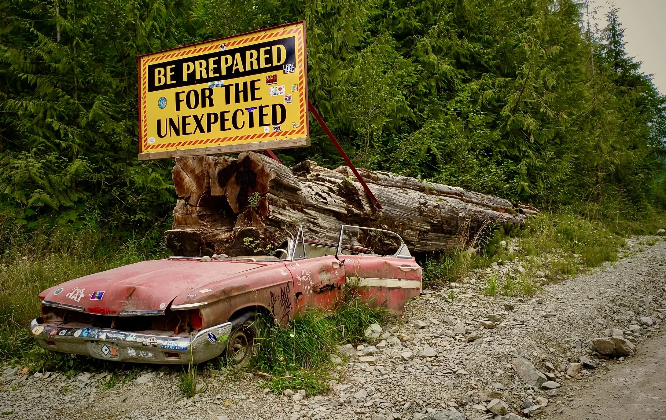 An old, rusted red convertible car next to a fallen tree trunk on a gravel roadside, with a yellow sign reading "Be Prepared for the Unexpected" in the background, surrounded by green trees.
