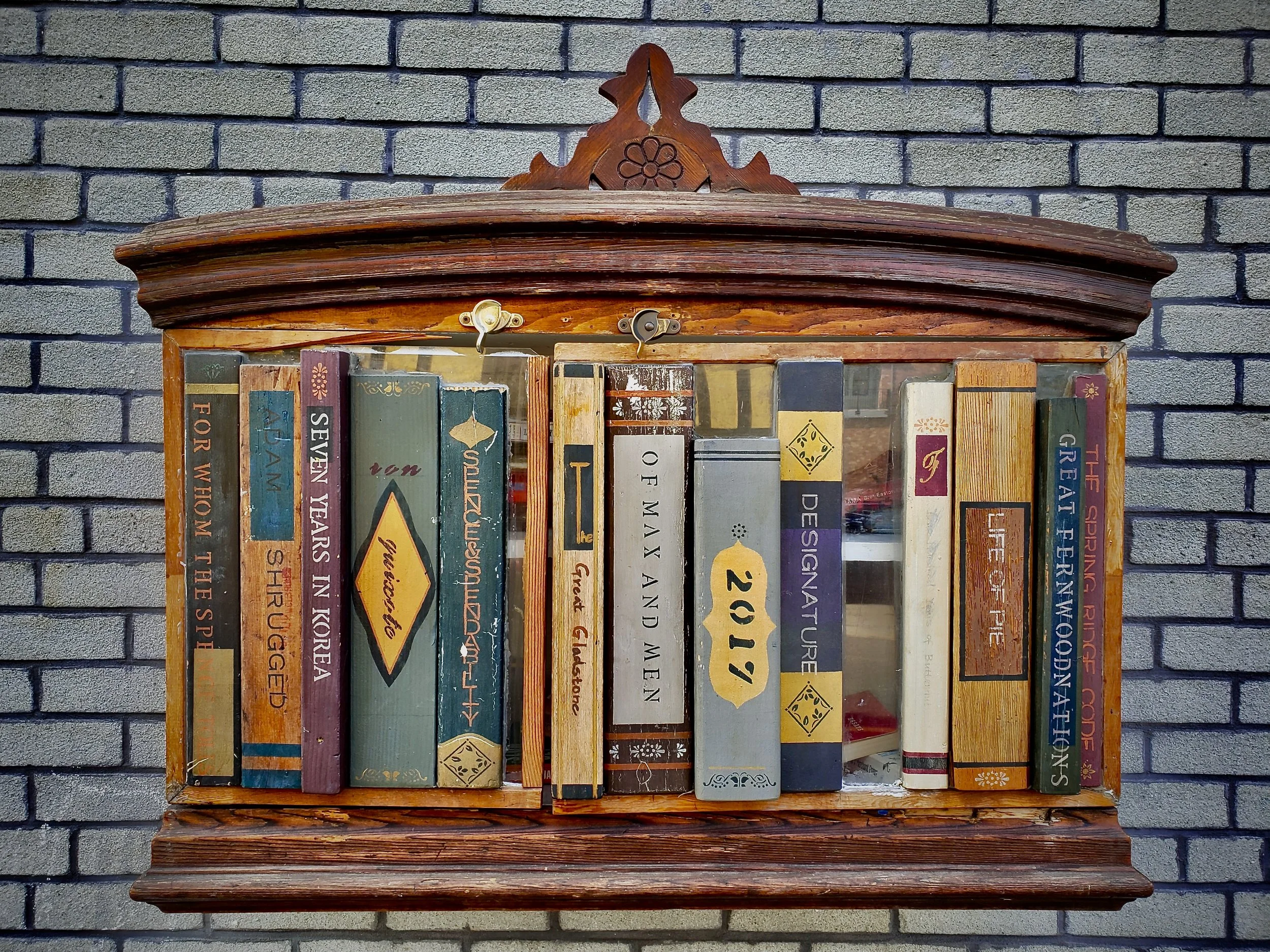 A wooden bookshelf mounted on a gray brick wall holding a collection of books.