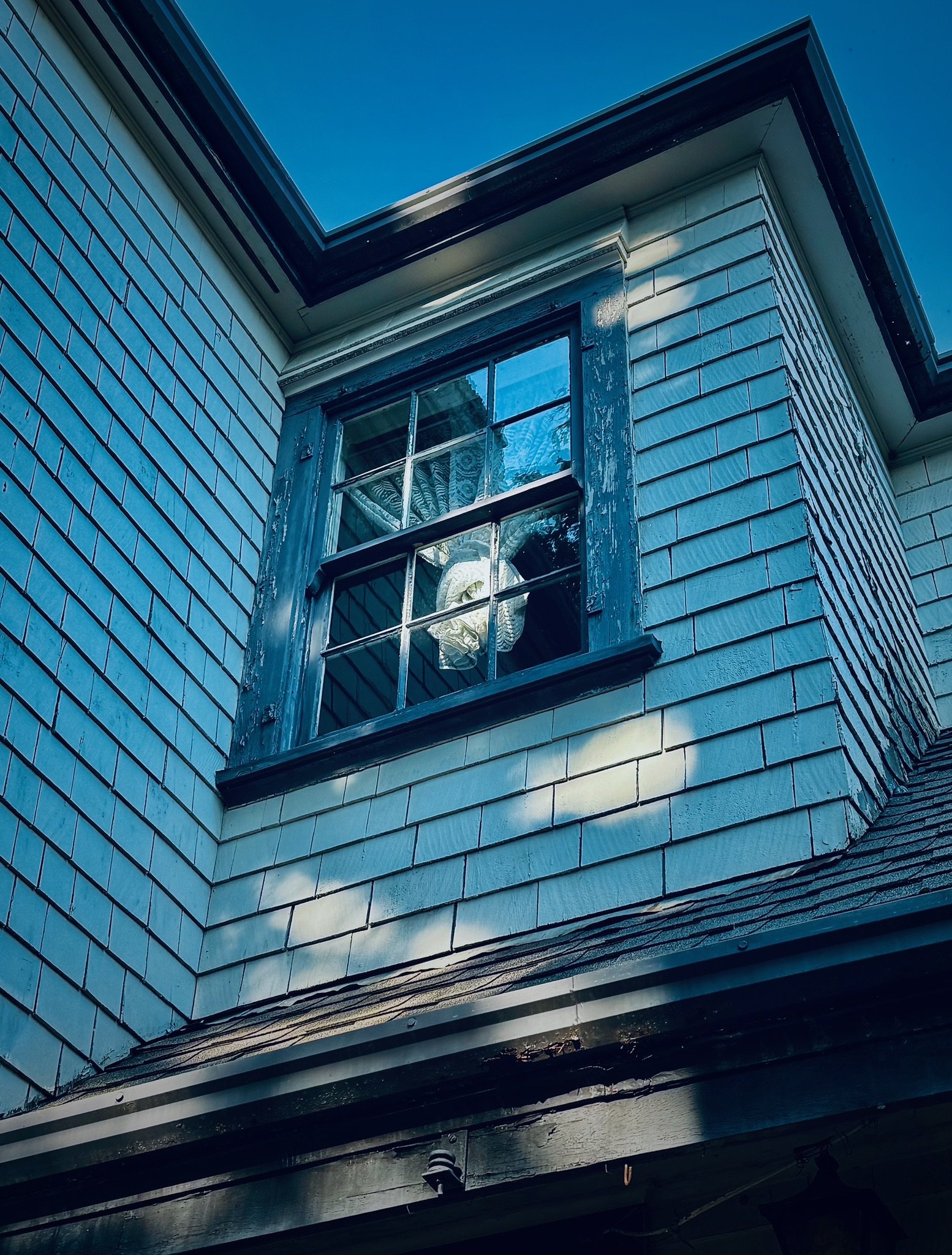 A close-up of a house's exterior corner showing a single window with peeling blue paint, partially open, revealing a curtain that looks like a skull inside. The house has blue siding and shingles, with a blue sky in the background.