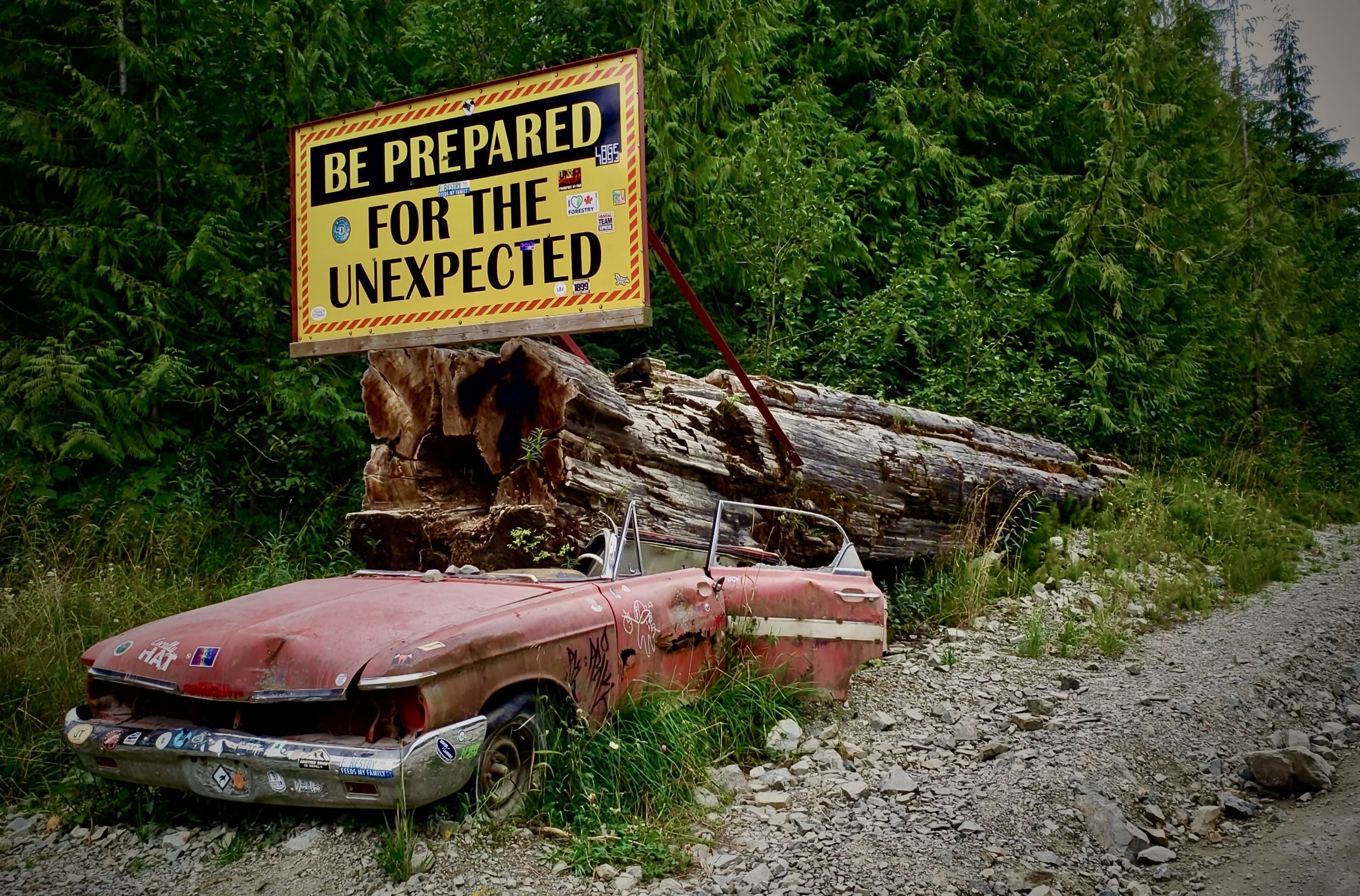 An old abandoned red car covered in stickers, parked beside a large fallen log and a yellow sign that reads, 'Be Prepared for the Unexpected', in a forest setting.