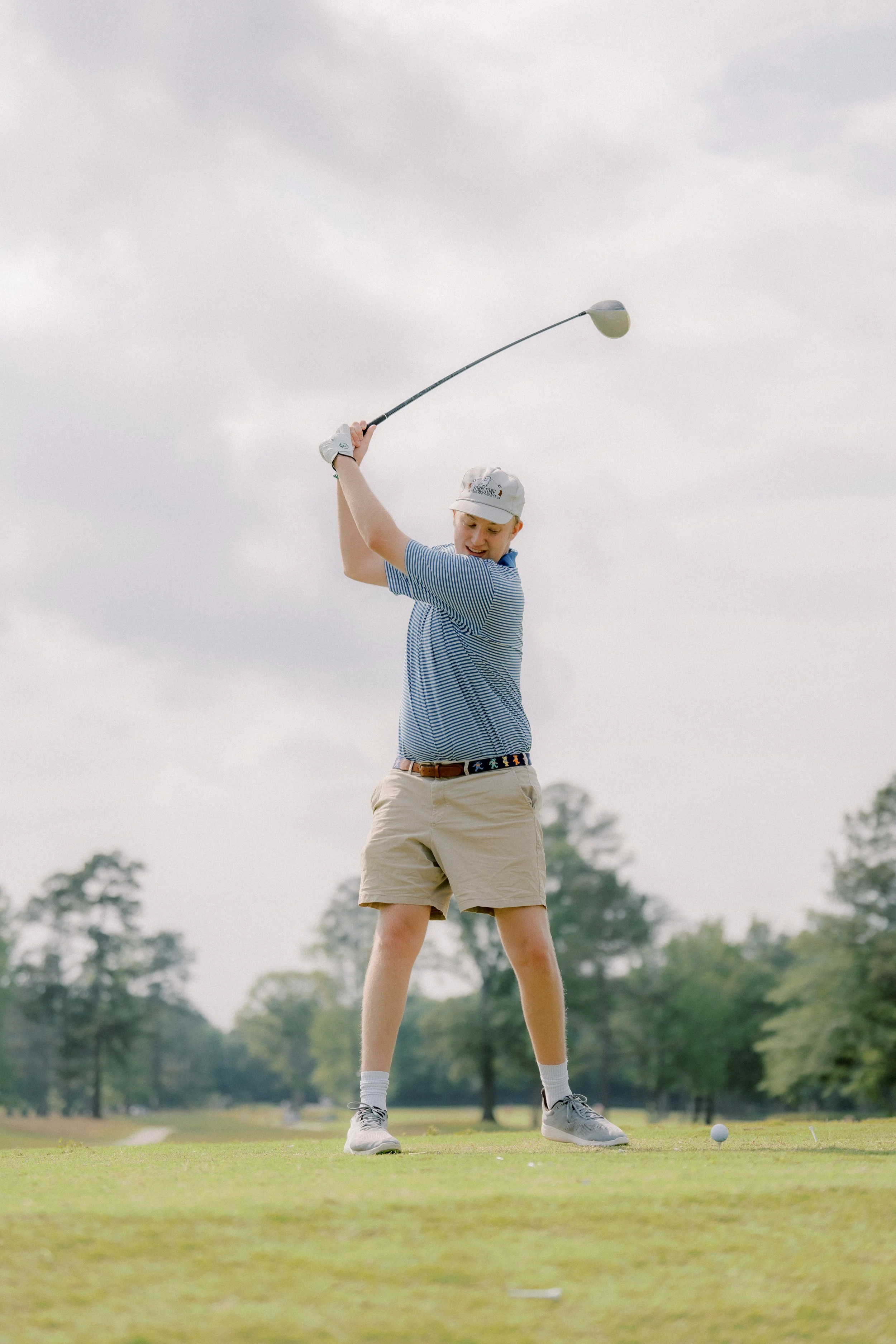 Young male golfer in striped shirt and beige shorts swinging a golf club on a golf course with trees in the background.