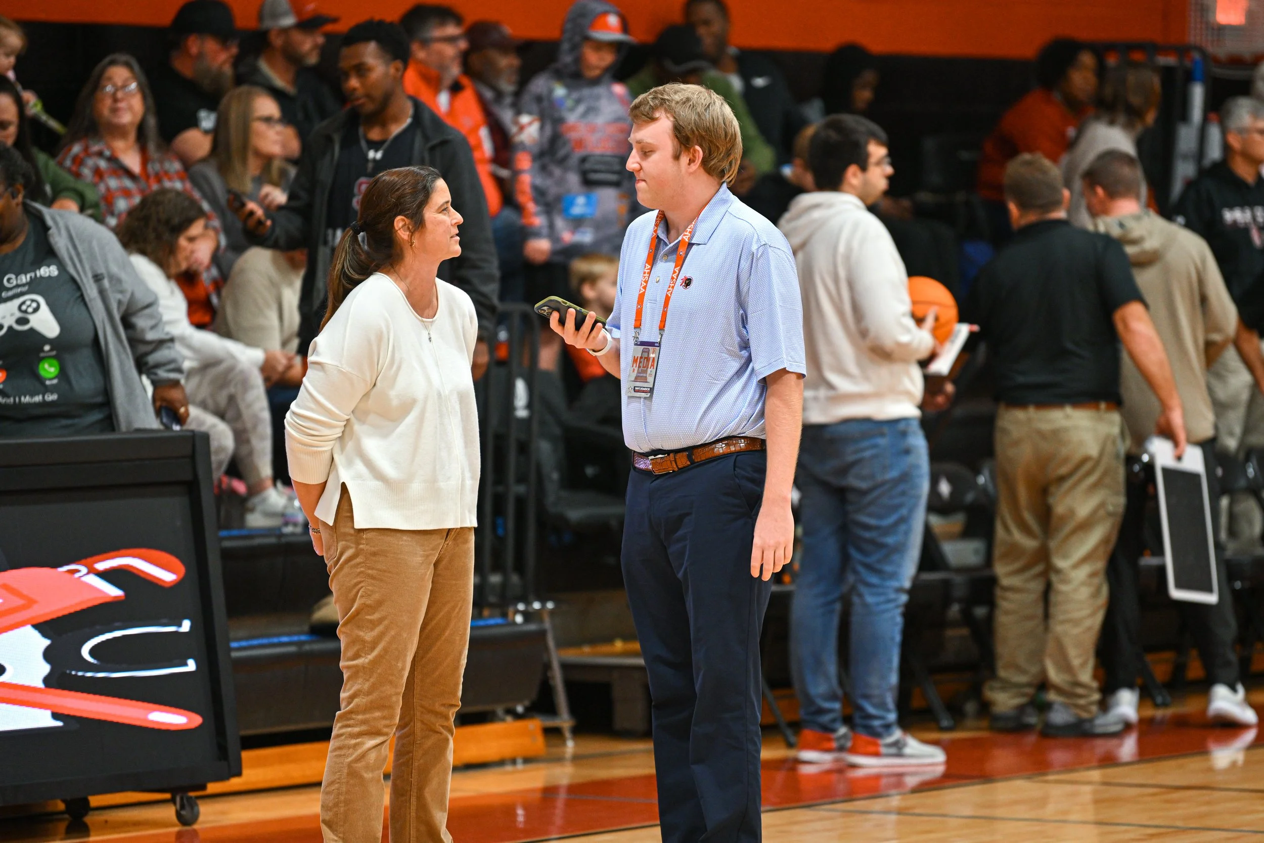 A woman and a man are talking on a basketball court during a game, with spectators seated in the background.