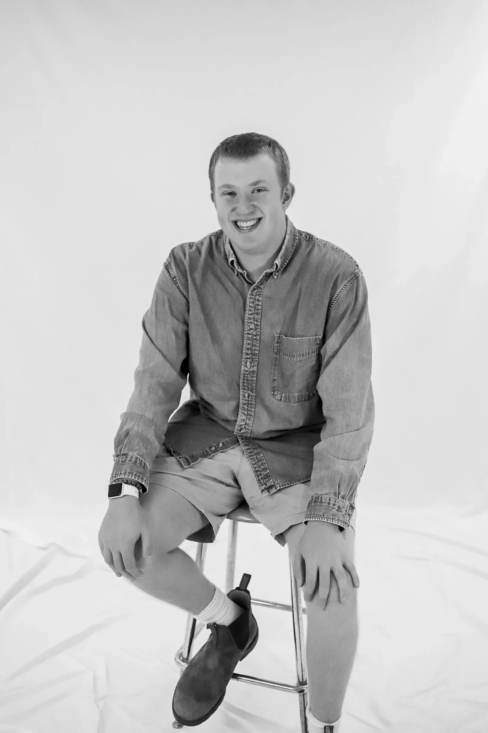 Smiling young man sitting on a stool against a plain background, wearing a denim shirt, shorts, socks, and boots.