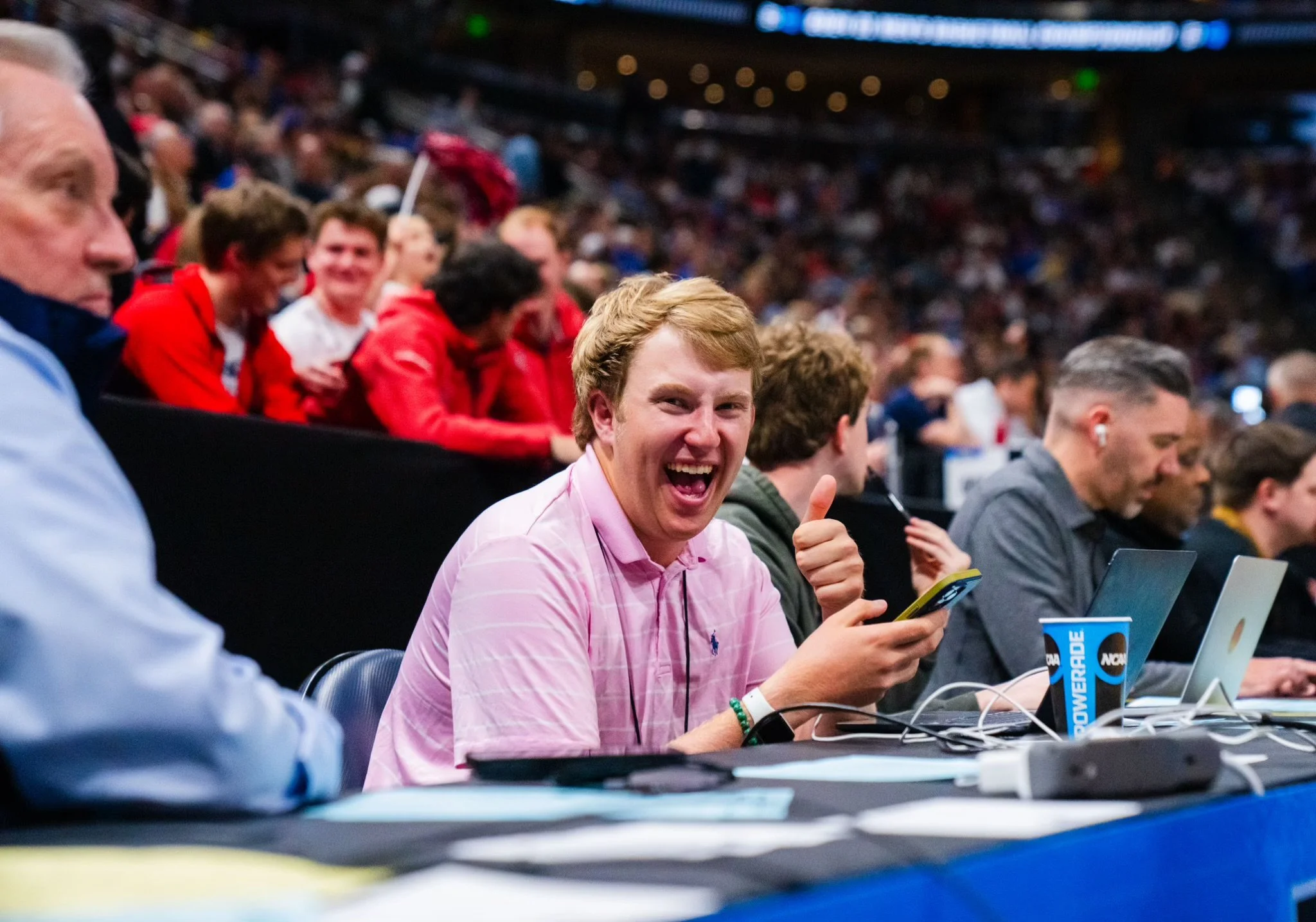 A young man in a pink shirt smiling, giving a thumbs-up while holding a phone, sitting at a table with laptops and a blue NCAA Gatorade cup, surrounded by crowd at an indoor event.