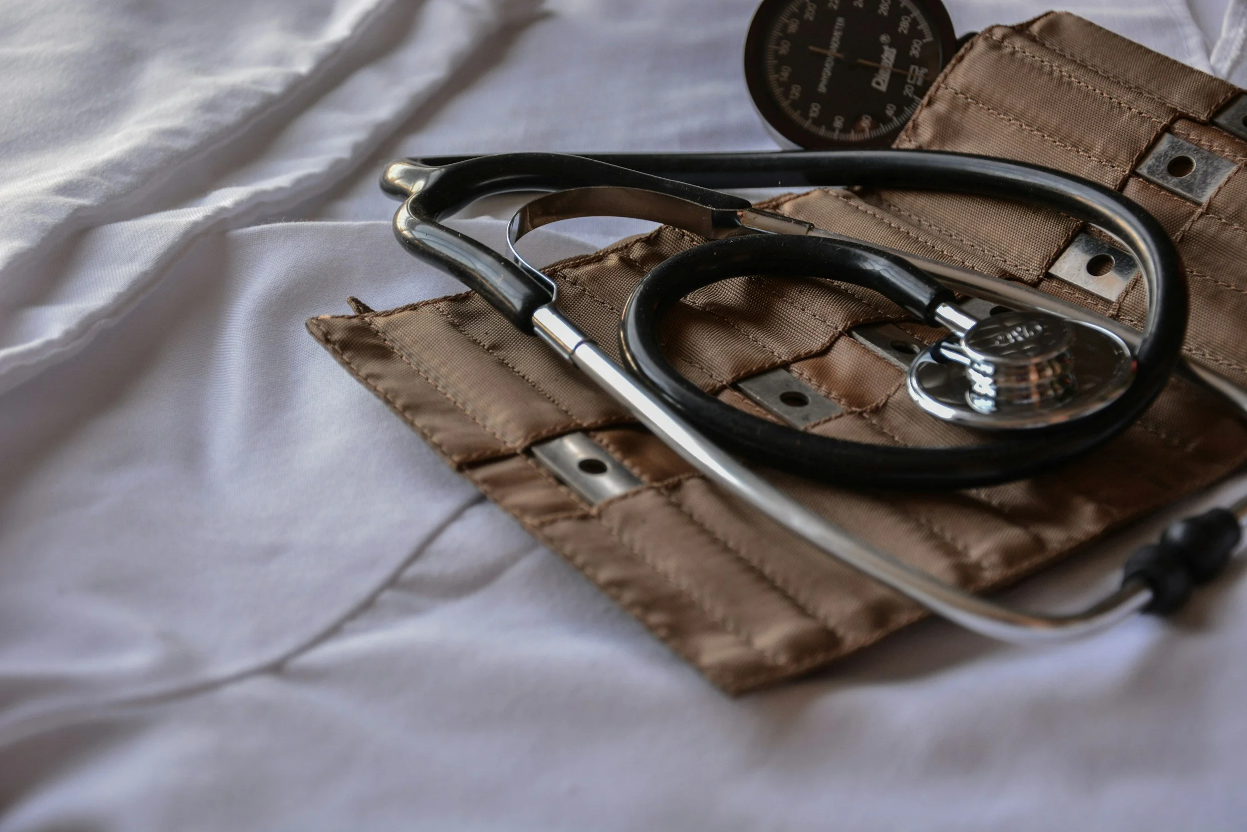 A stethoscope resting on a brown medical case with a black sphygmomanometer in the background on a white fabric surface.