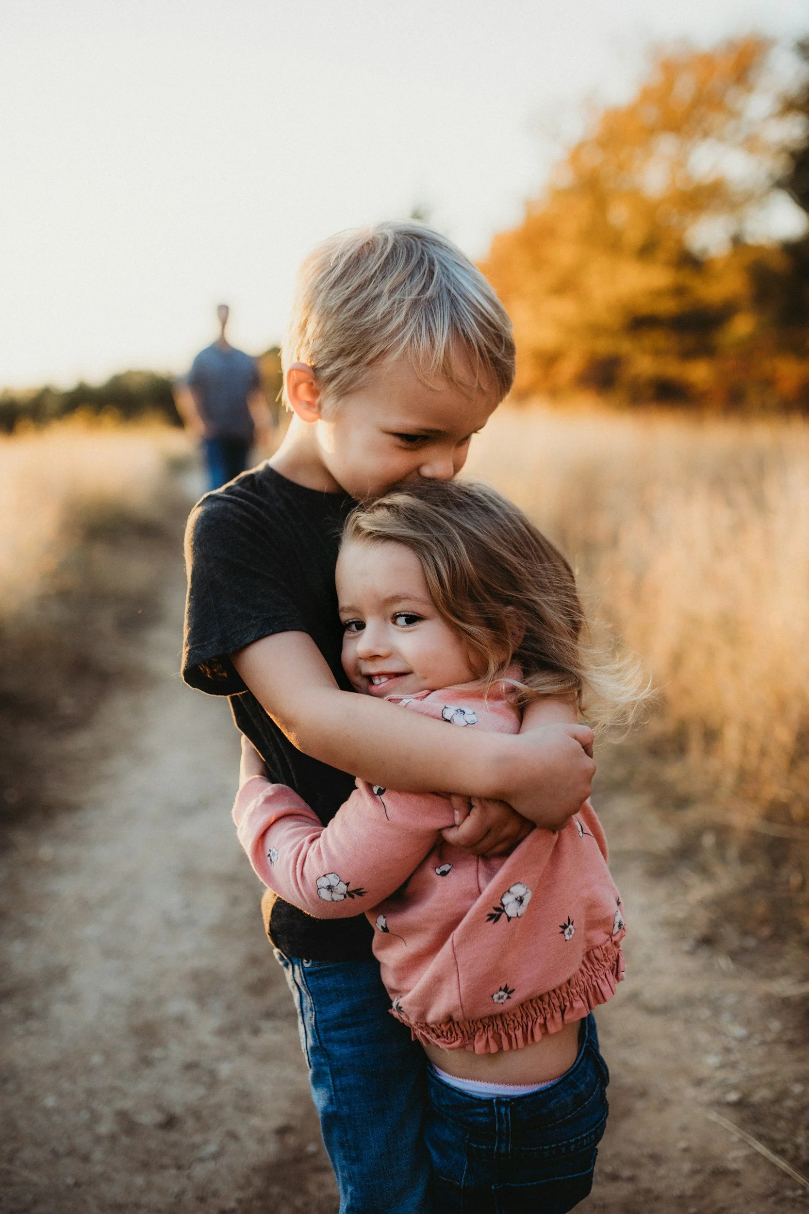 Two children hugging on a dirt path outdoors during sunset, with a man in the background and fall-colored trees.