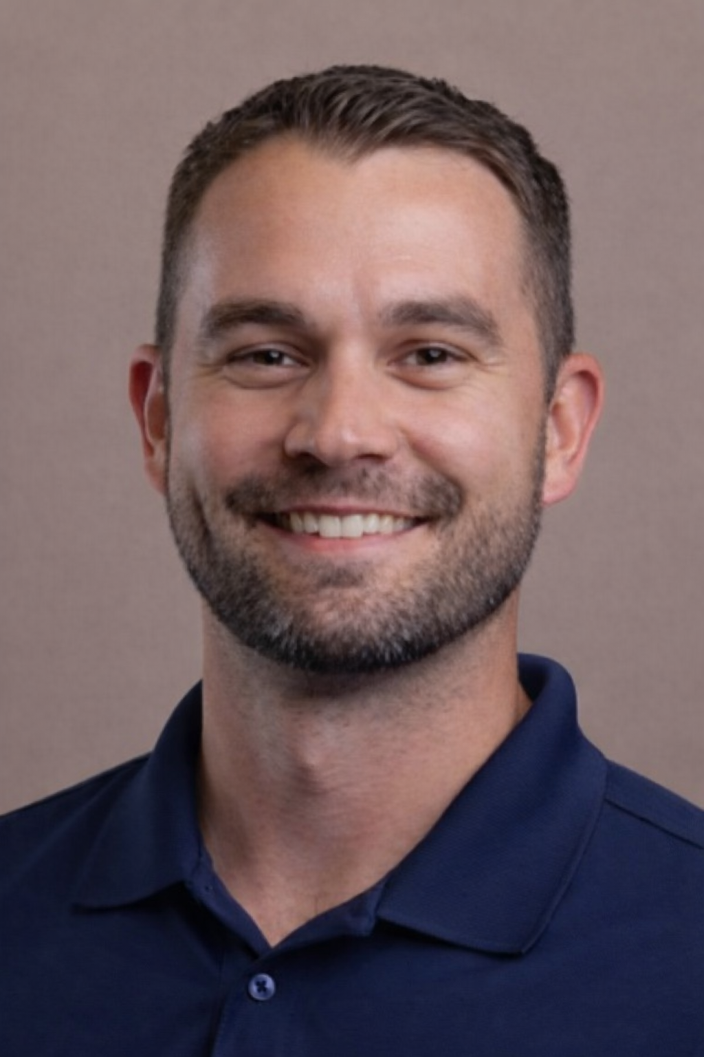Smiling man with short hair and beard wearing a navy blue collared shirt, against a plain background.