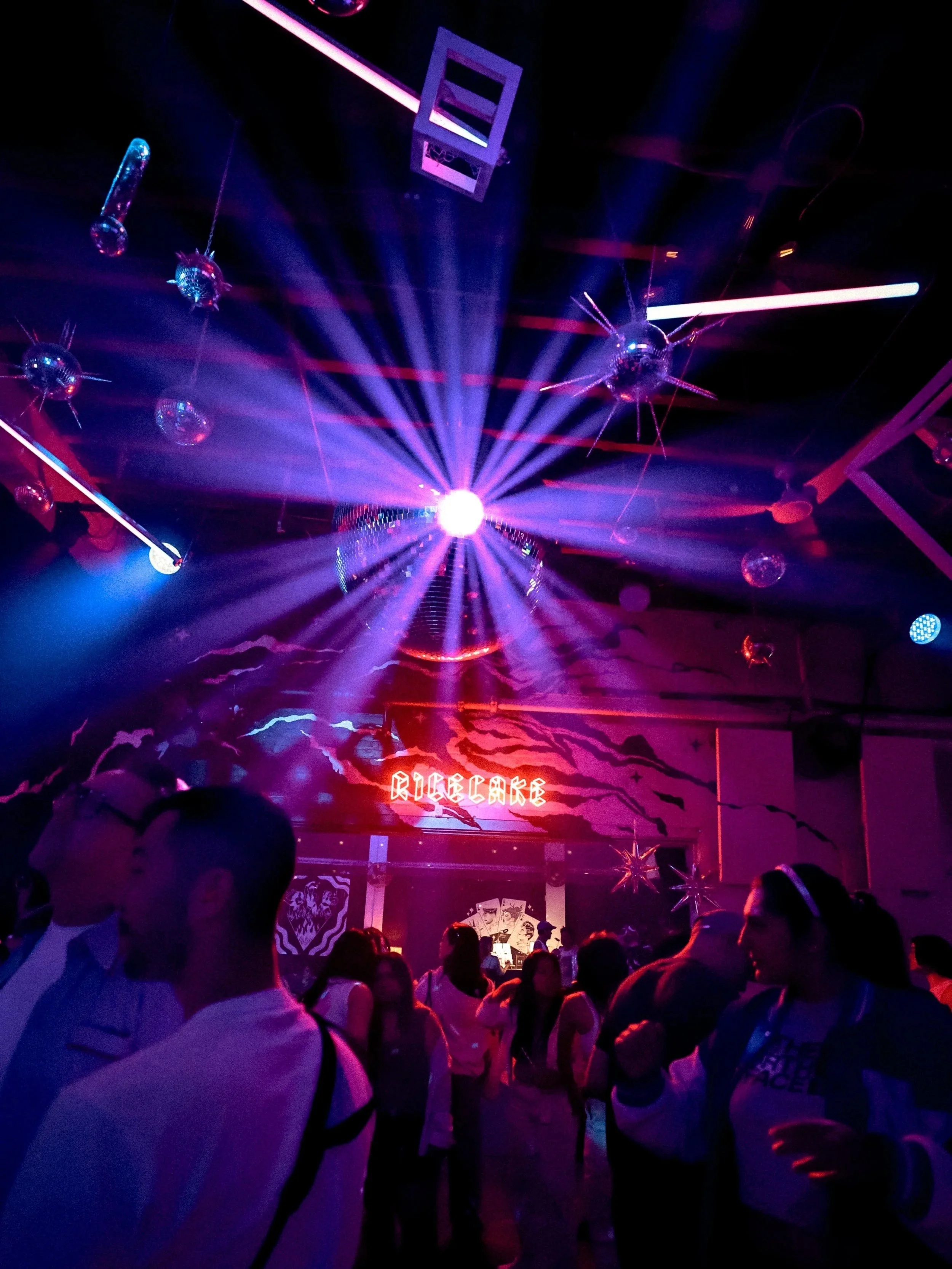A dimly lit nightclub scene with people dancing under colorful laser lights. Discos and decorative lighting hang from the ceiling, with a bright sign that reads 'RICE CAKE' in neon lights. The crowd appears to be enjoying the energetic atmosphere.