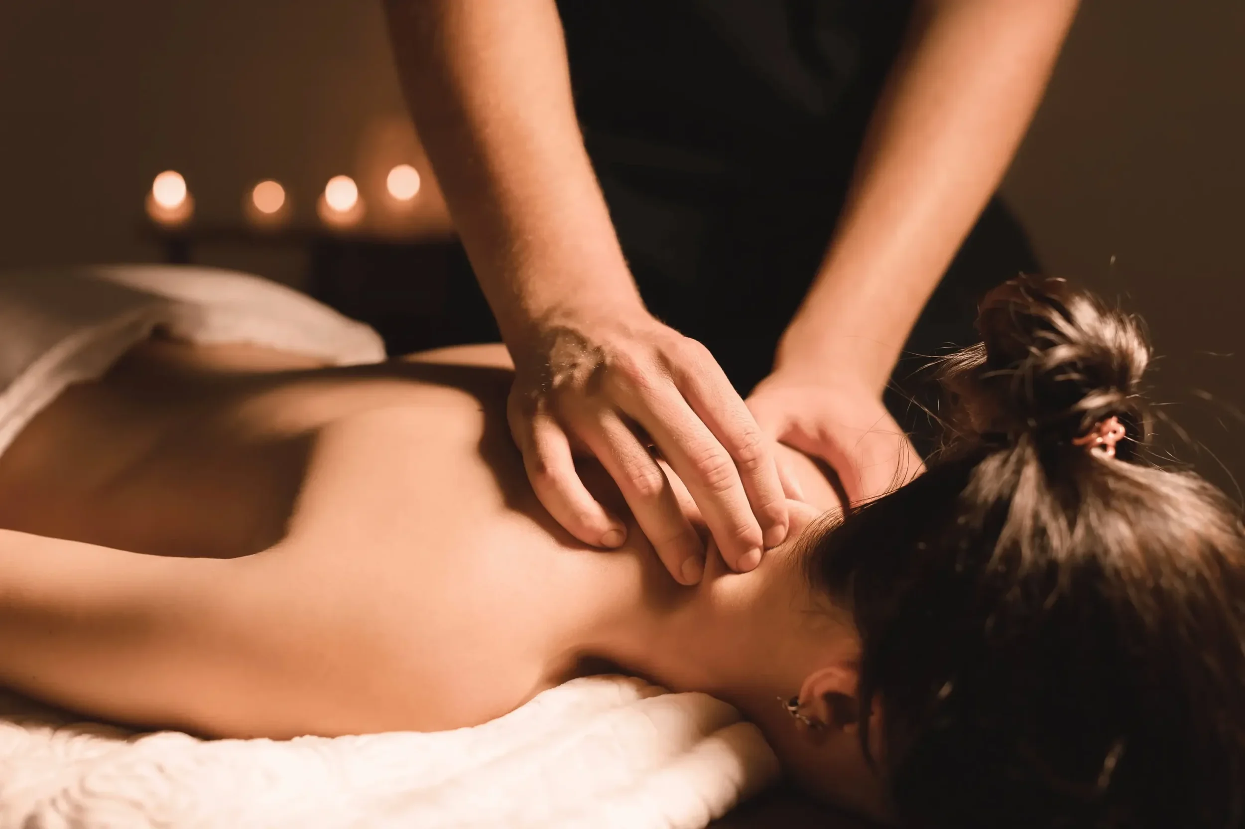A person receives a massage on their back while lying face down on a massage table with a white towel. Candles are visible in the background, creating a relaxing atmosphere.