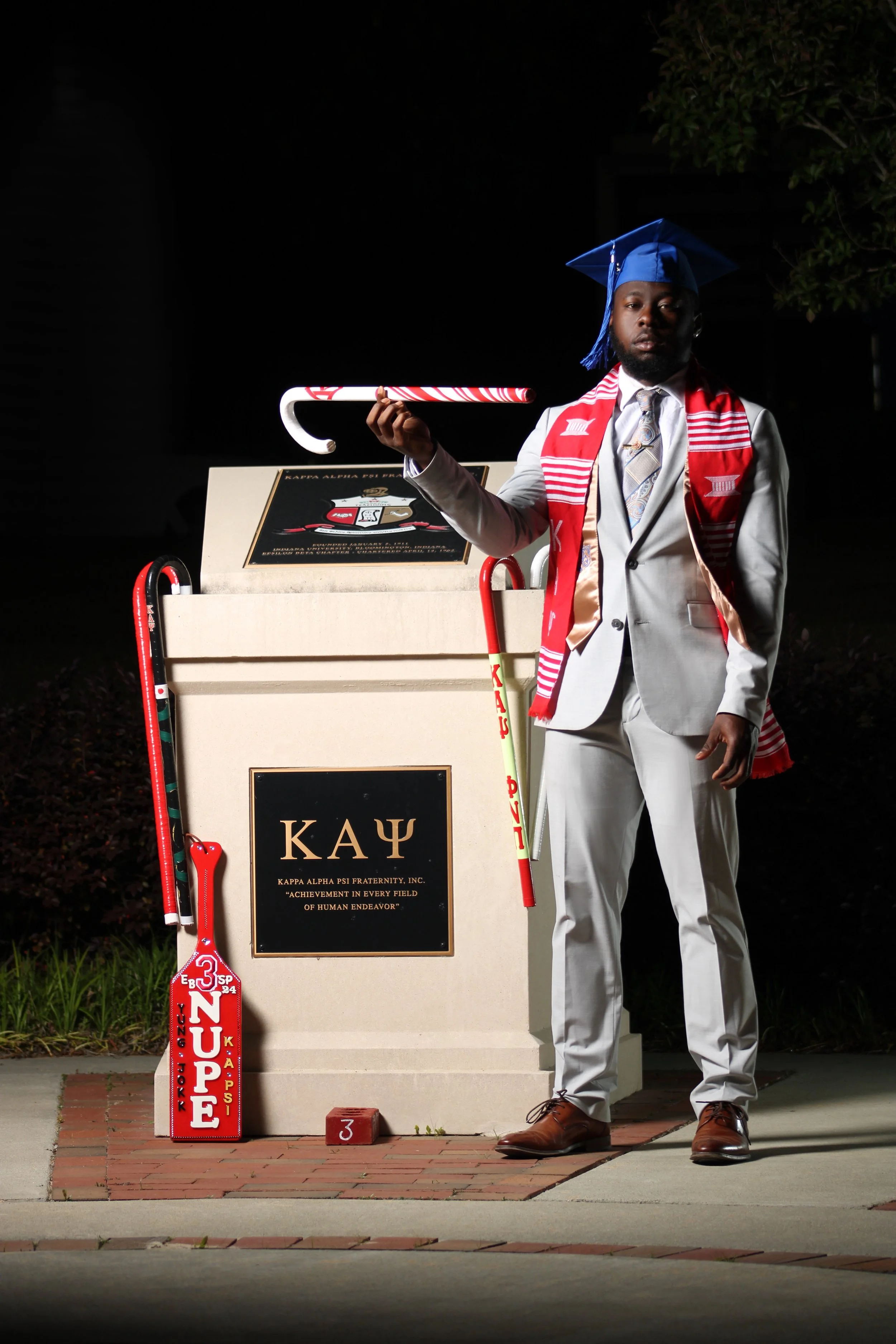 A young man in a gray suit and blue graduation cap stands next to a stone monument with a plaque for Kappa Alpha Psi Fraternity holding a candy cane. He is also wearing a red and white scarf and tie, and holds a cane with a red and white pattern. A r