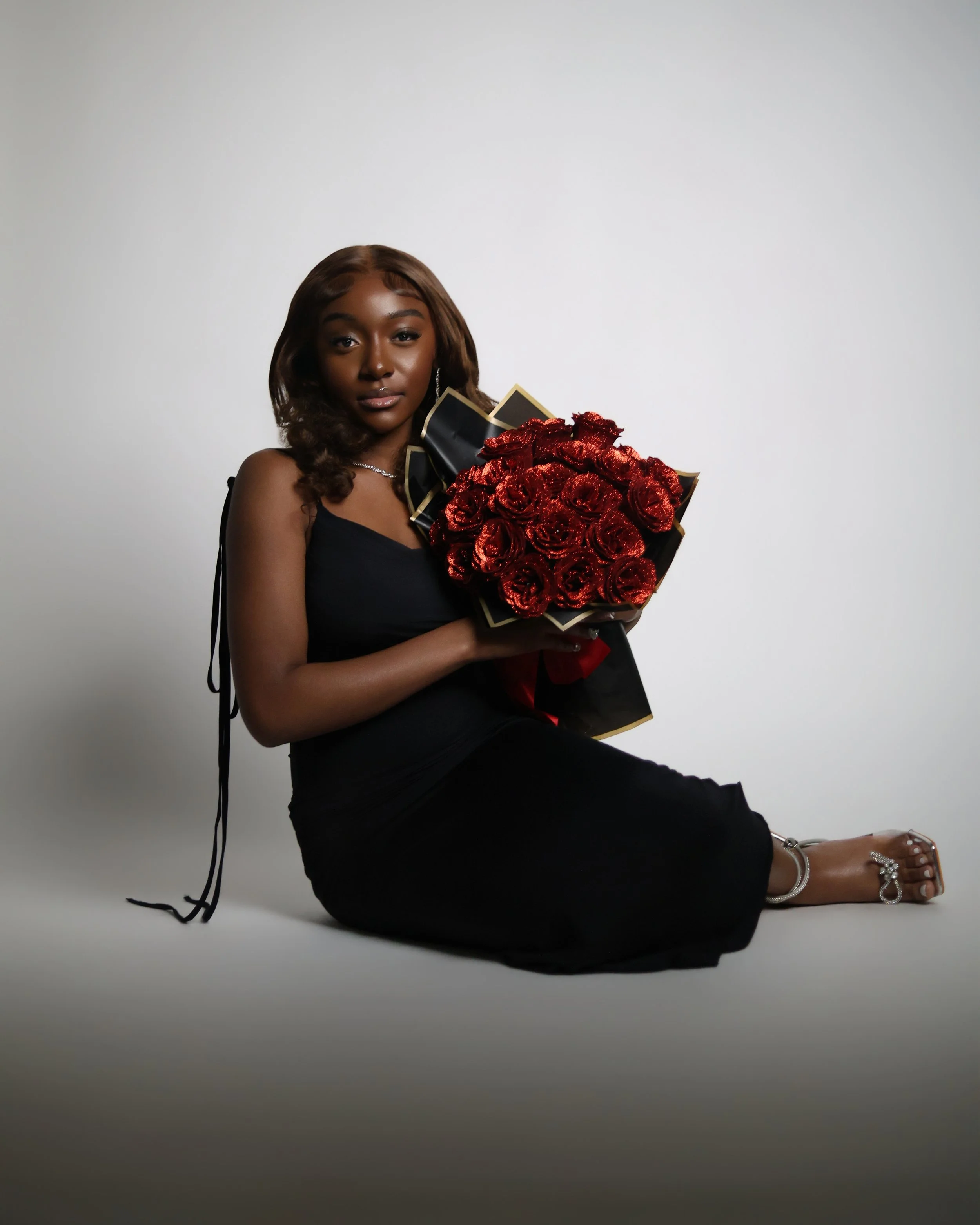 A woman in a black dress sitting on the floor, holding a bouquet of red roses, with a neutral white background.