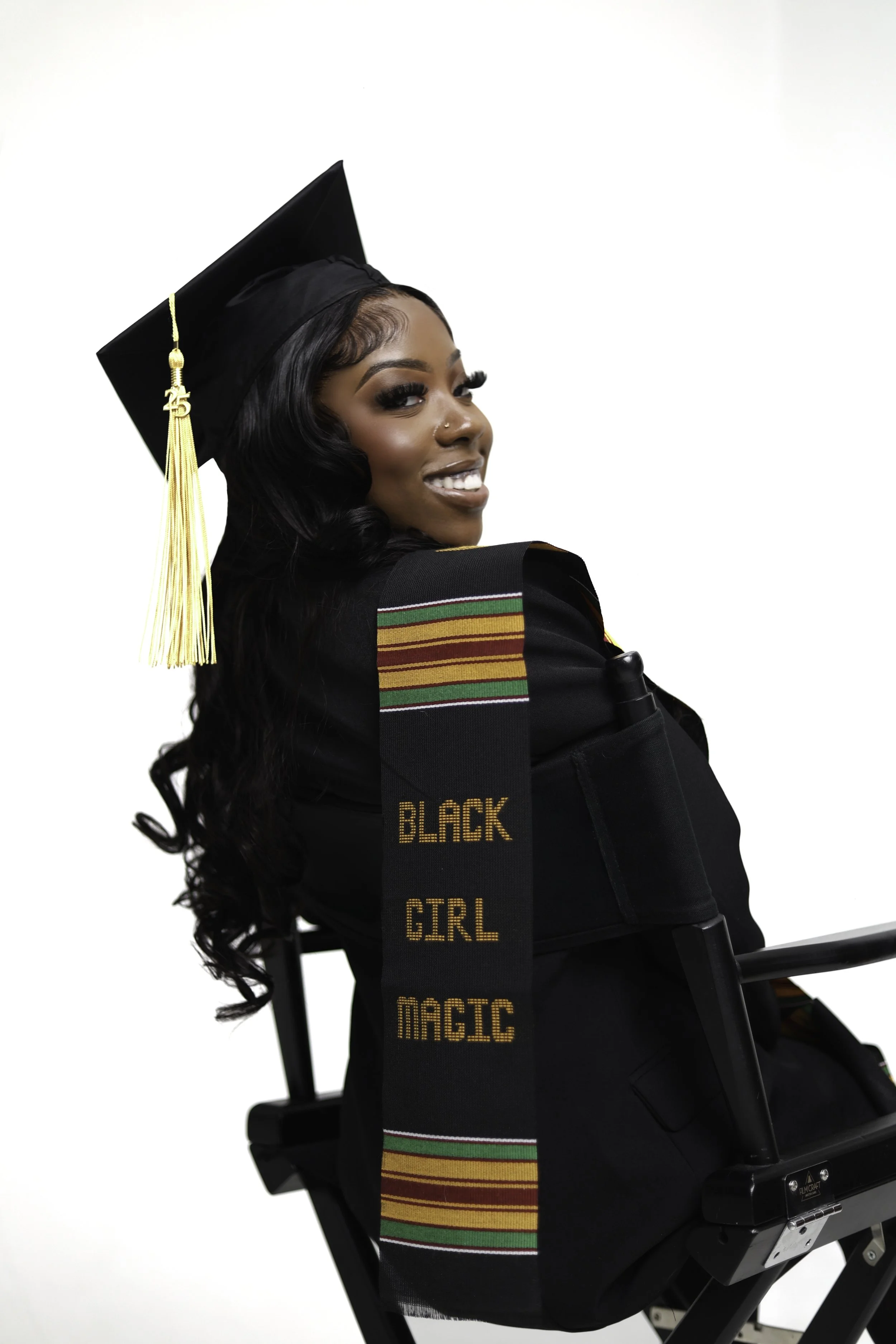 A young Black woman in a graduation cap and gown, smiling and sitting in a wheelchair. She wears a sash with colorful stripes and the words "Black Girl Magic."