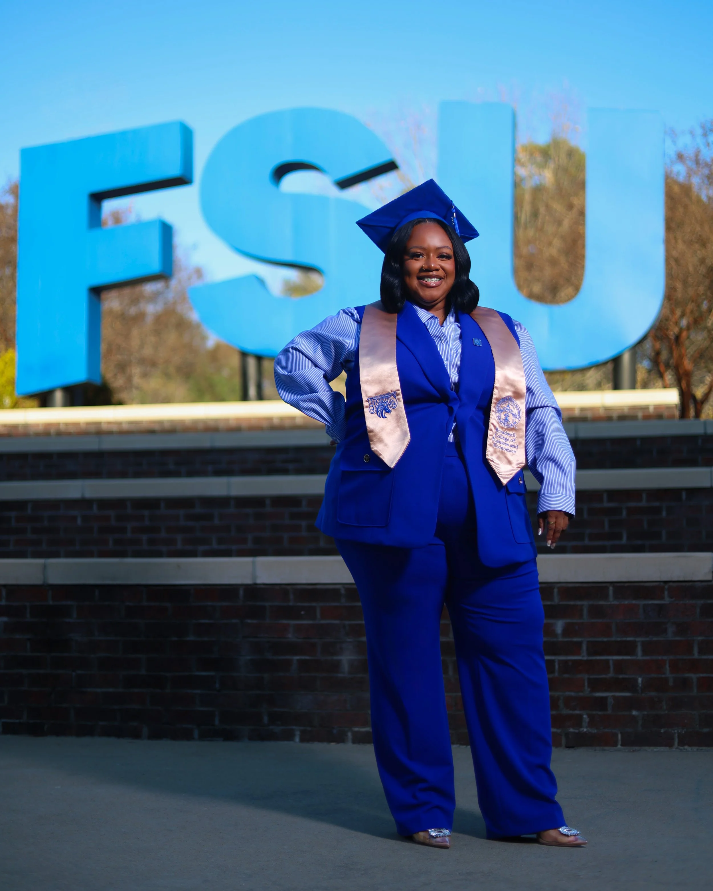 A woman in a blue graduation cap and gown standing in front of large blue letters spelling 'FSU'. She is smiling and wearing a sash and heels.