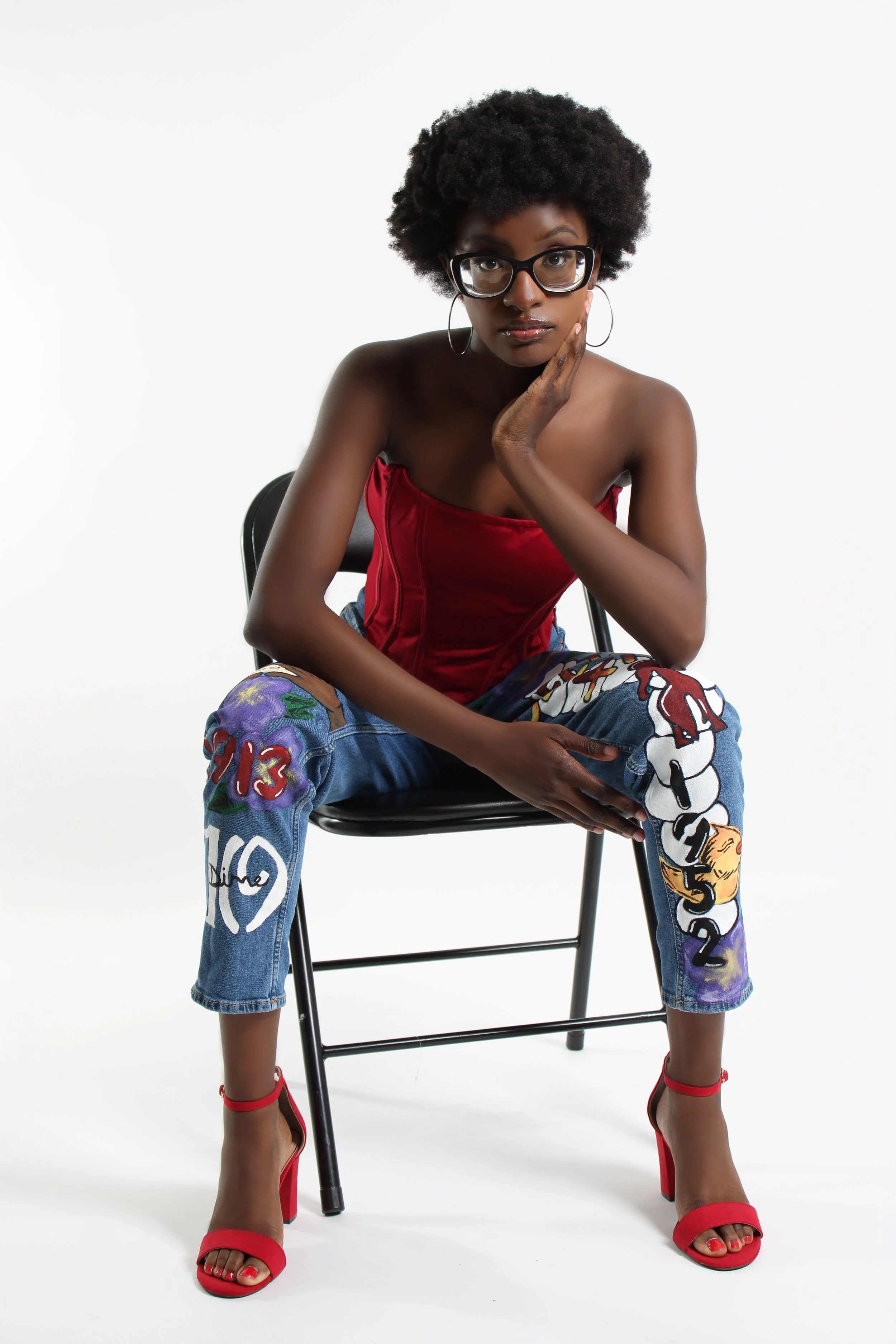 A woman with curly hair wearing glasses, a red strapless top, colorful graffiti-style jeans, and red high-heeled shoes, sitting on a black chair against a white background.