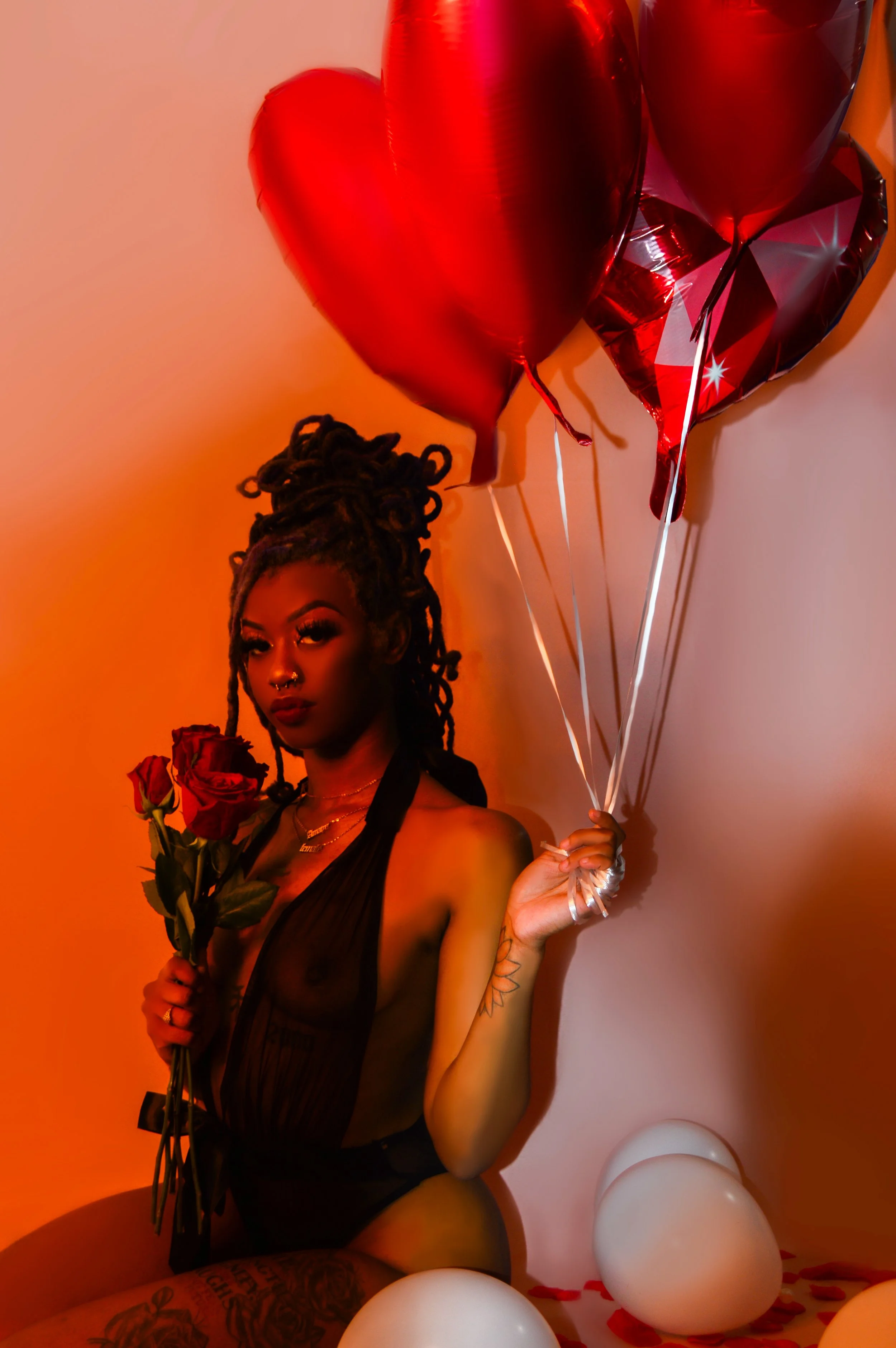 A woman with dreadlocks holding red roses and heart-shaped balloons, dressed in black sheer clothing, with tattoos and jewelry, surrounded by white balloons on the floor.