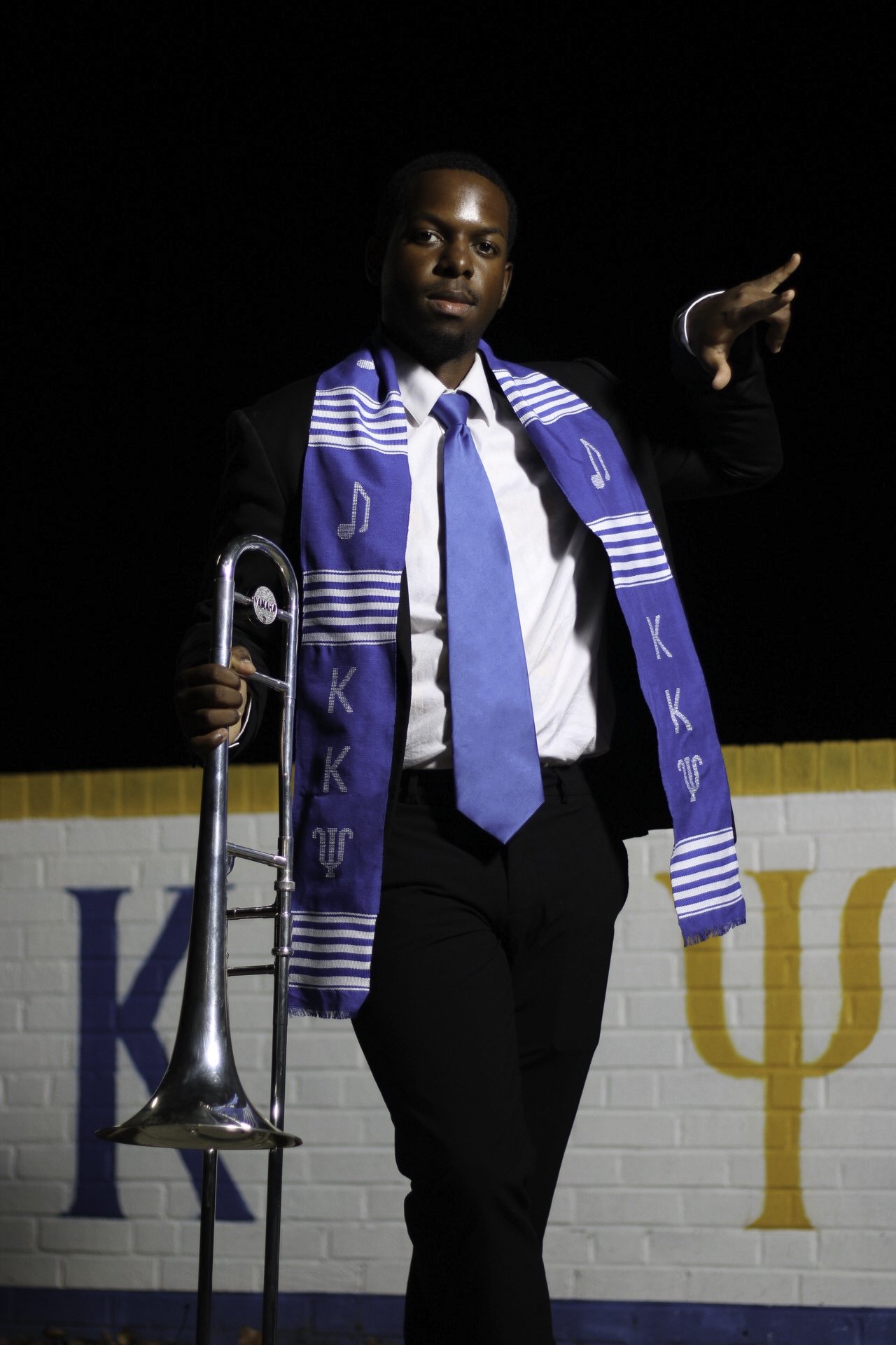 A man in a suit holding a trombone and wearing a blue sorority stole stands in front of a white wall with yellow borders and Greek letters 'K' and 'Y' painted on it.