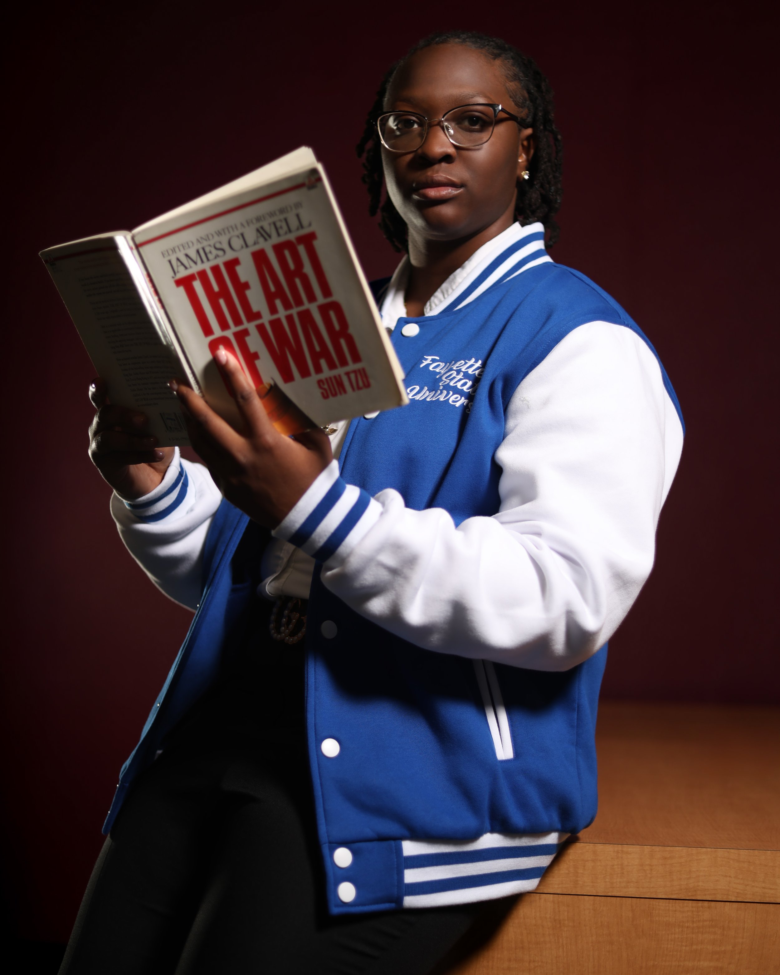 A woman wearing glasses and a blue and white varsity jacket reading a book titled "The Art of War" by Sun Tzu against a dark background.