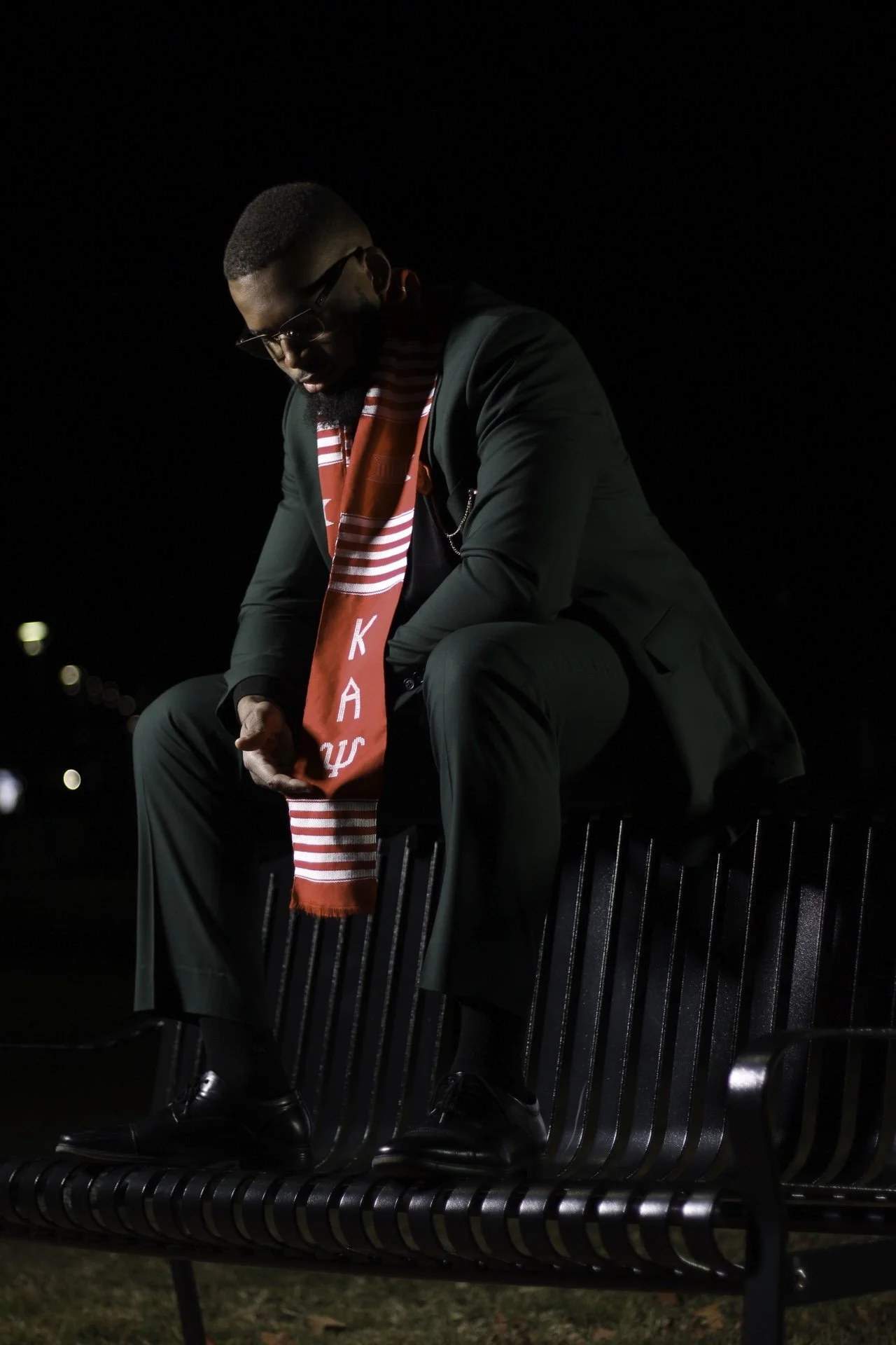 A man dressed in a dark green suit, black shoes, glasses, and a red and white Sports team scarf sitting on a black metal bench at night, with his head bowed.