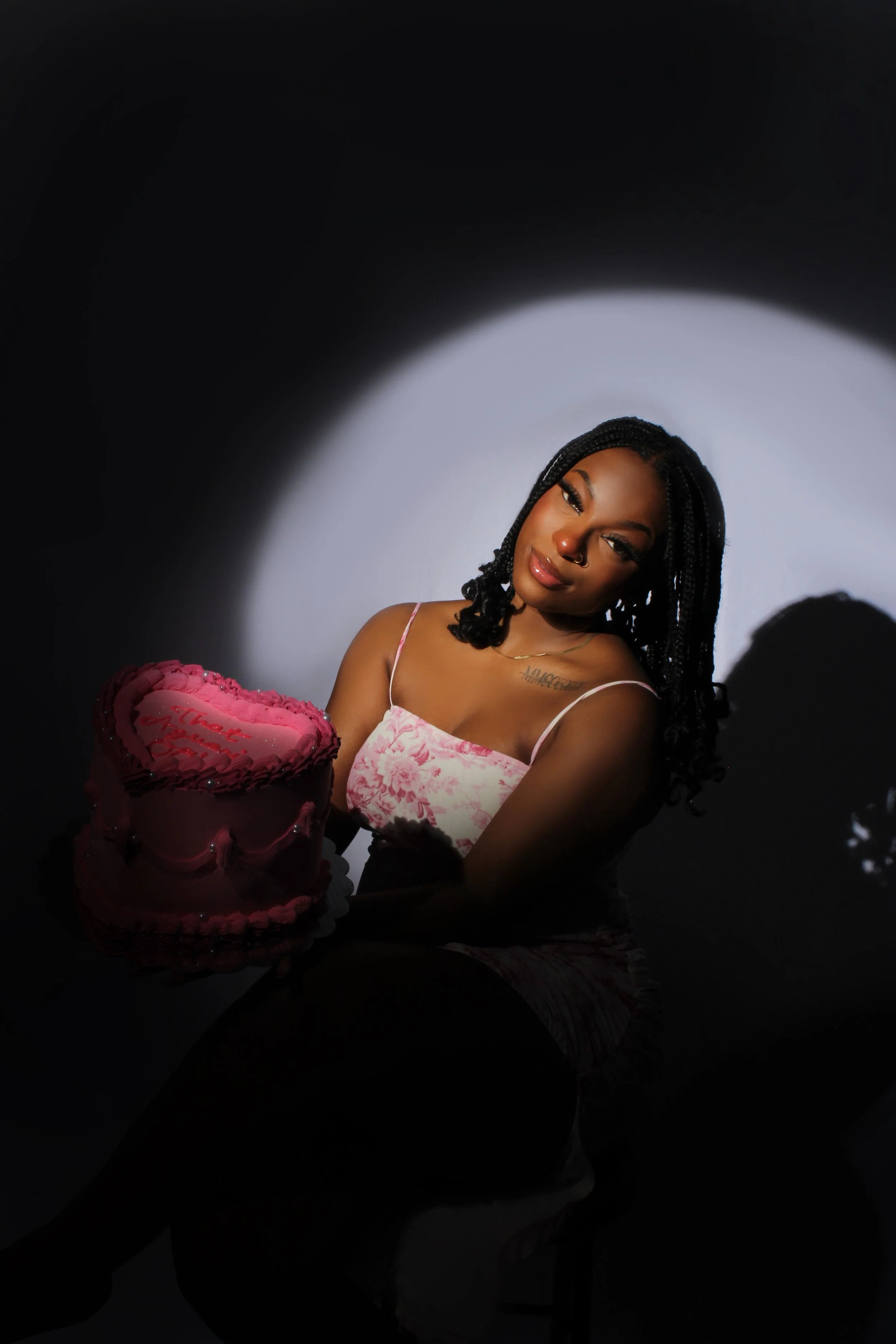 Woman with braided hair holding a pink heart-shaped cake with red writing, illuminated by a spotlight against a dark background.