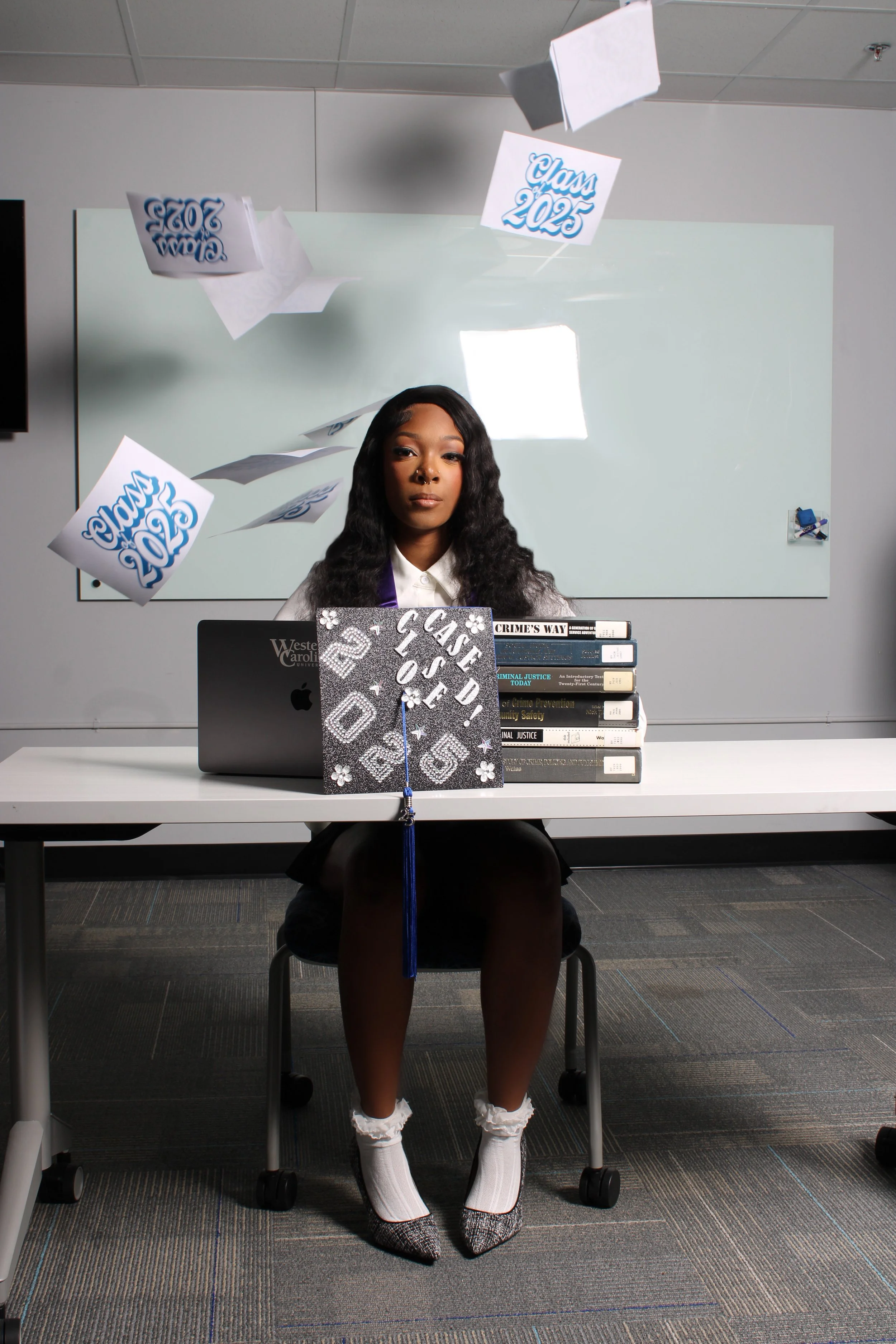 A young woman in a black skirt and lace heels with white socks sits at a desk decorated for graduation, with a dove-shaped cap in front and a black MacBook laptop. A graduation cap and waist-high stacks of law books are on the desk, and graduation ba