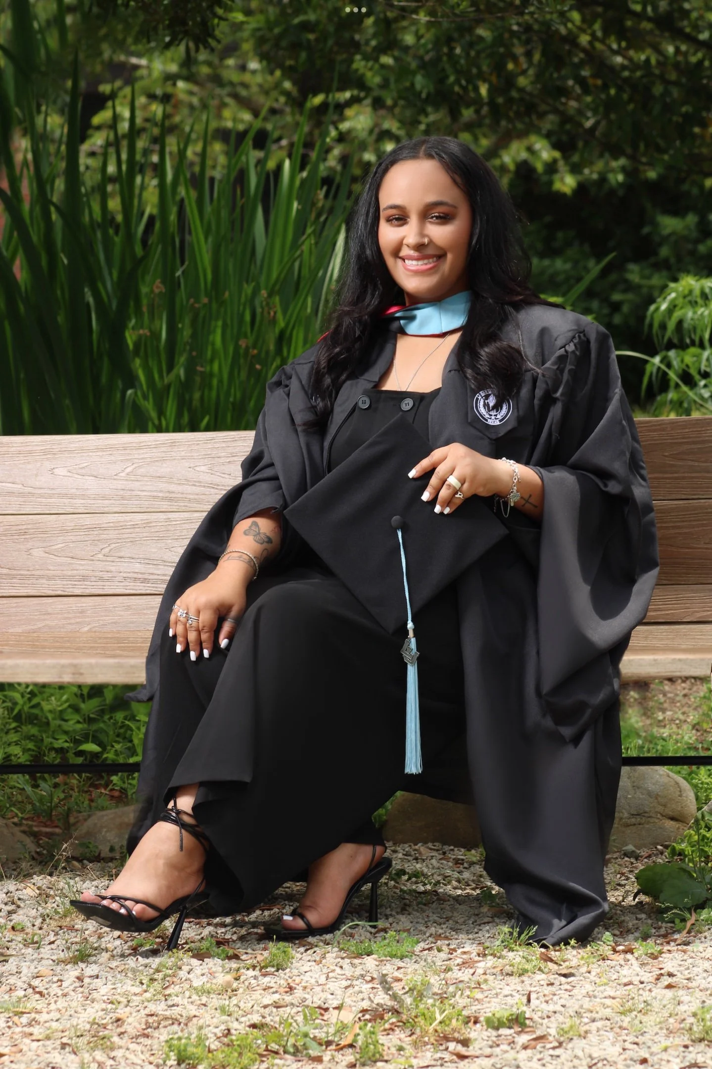A woman in a graduation gown and cap sitting on a wooden bench outdoors, holding her cap, with greenery in the background.