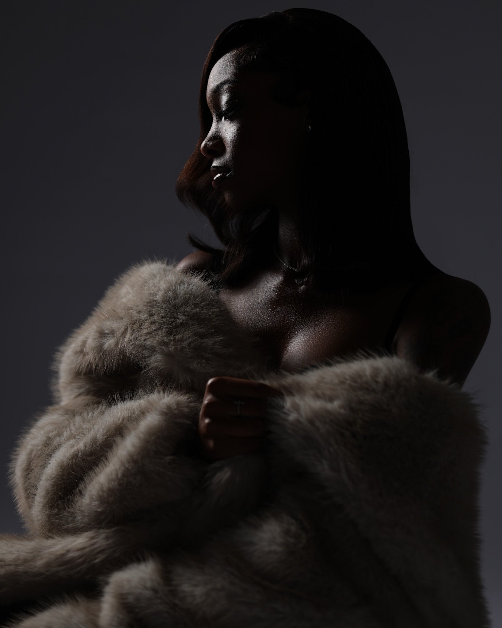 A woman with dark skin and shoulder-length hair wearing a fur coat, shown in profile against a dark background.