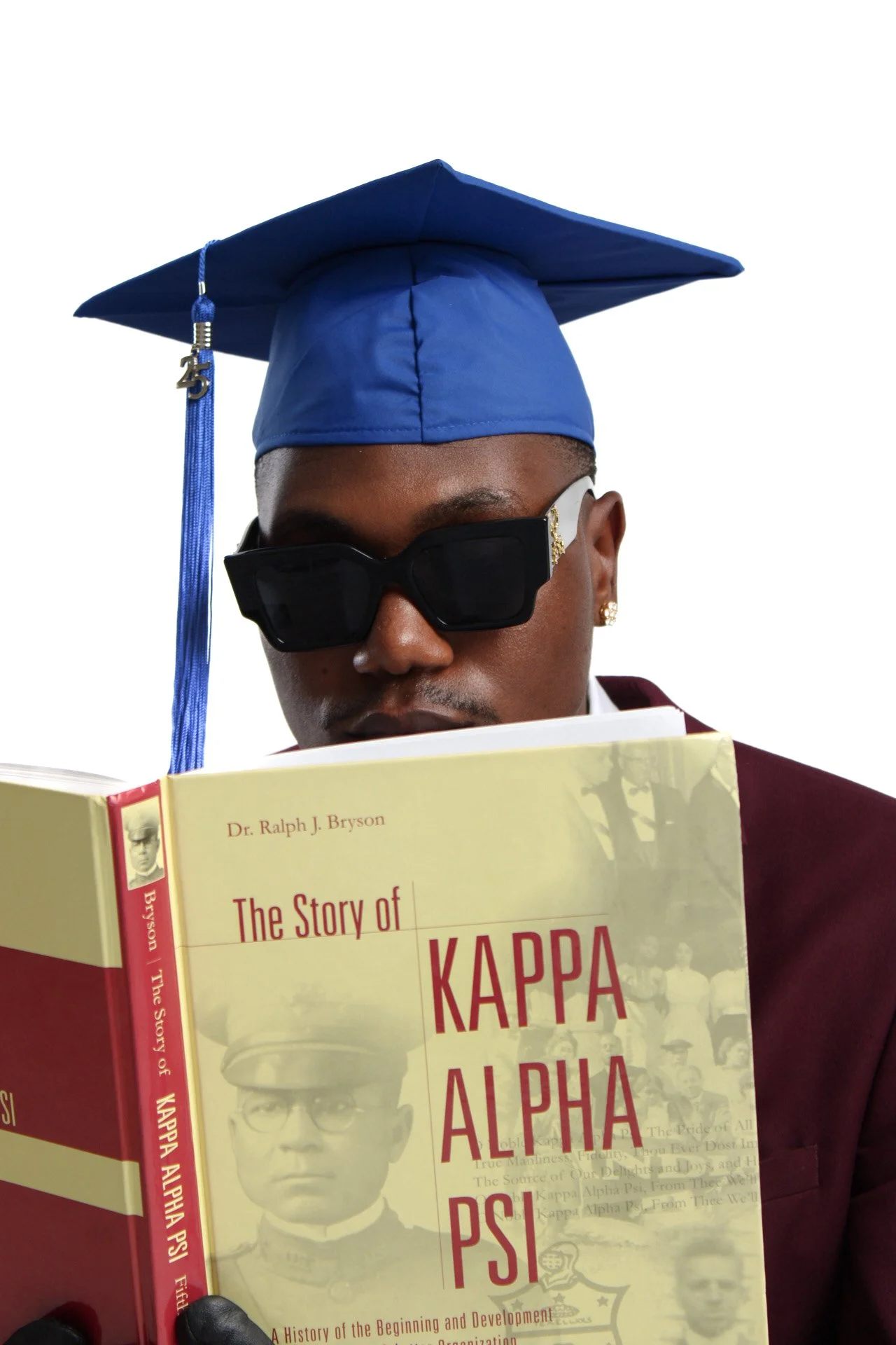 A young man wearing a blue graduation cap and sunglasses, holding a book titled 'The Story of Kappa Alpha Psi' by Dr. Ralph J. Bryson.