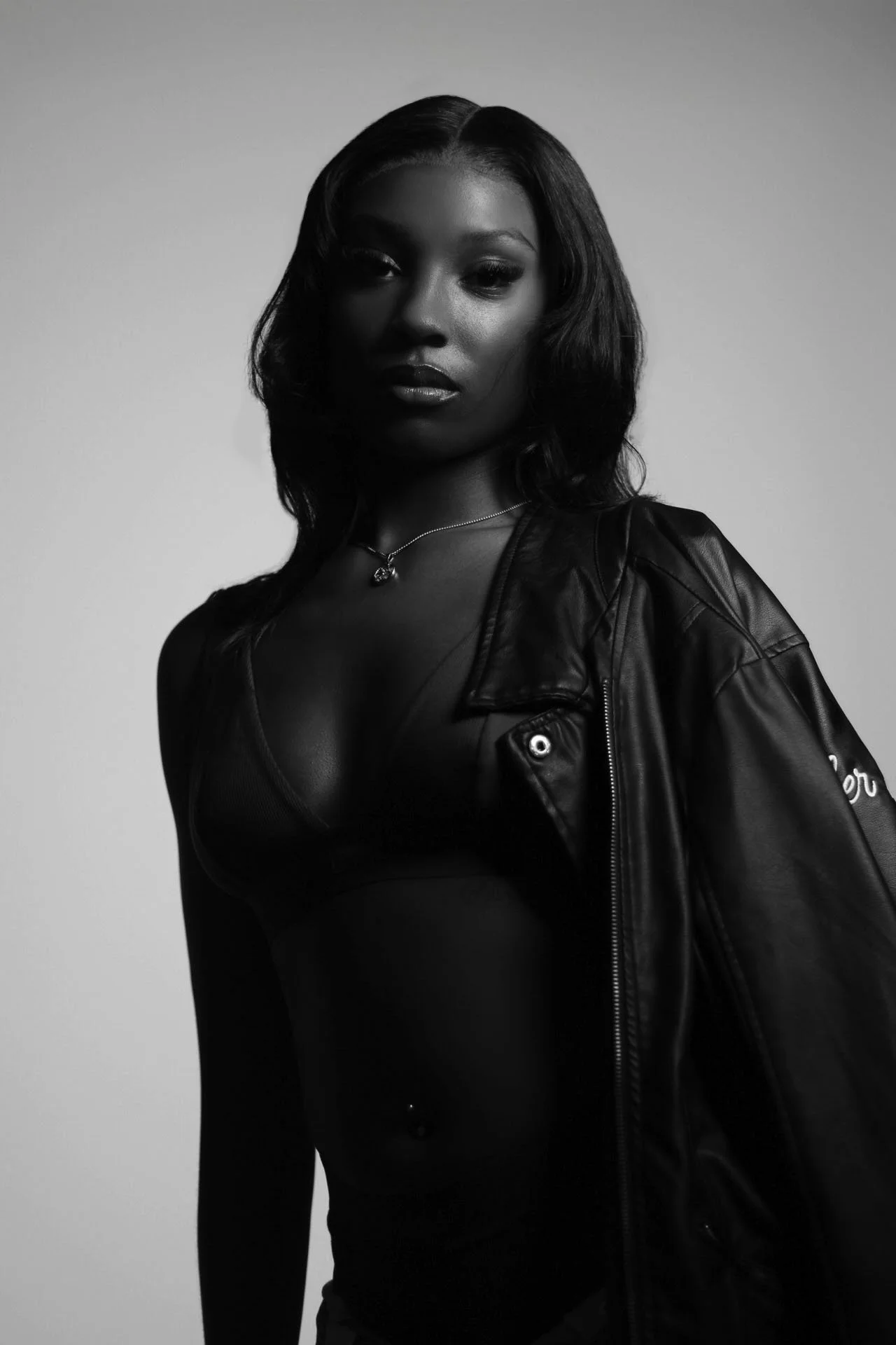 Black and white photo of a woman with shoulder-length hair, wearing a leather jacket draped over her shoulder, a necklace, and a sheer top with a piercing on her stomach.