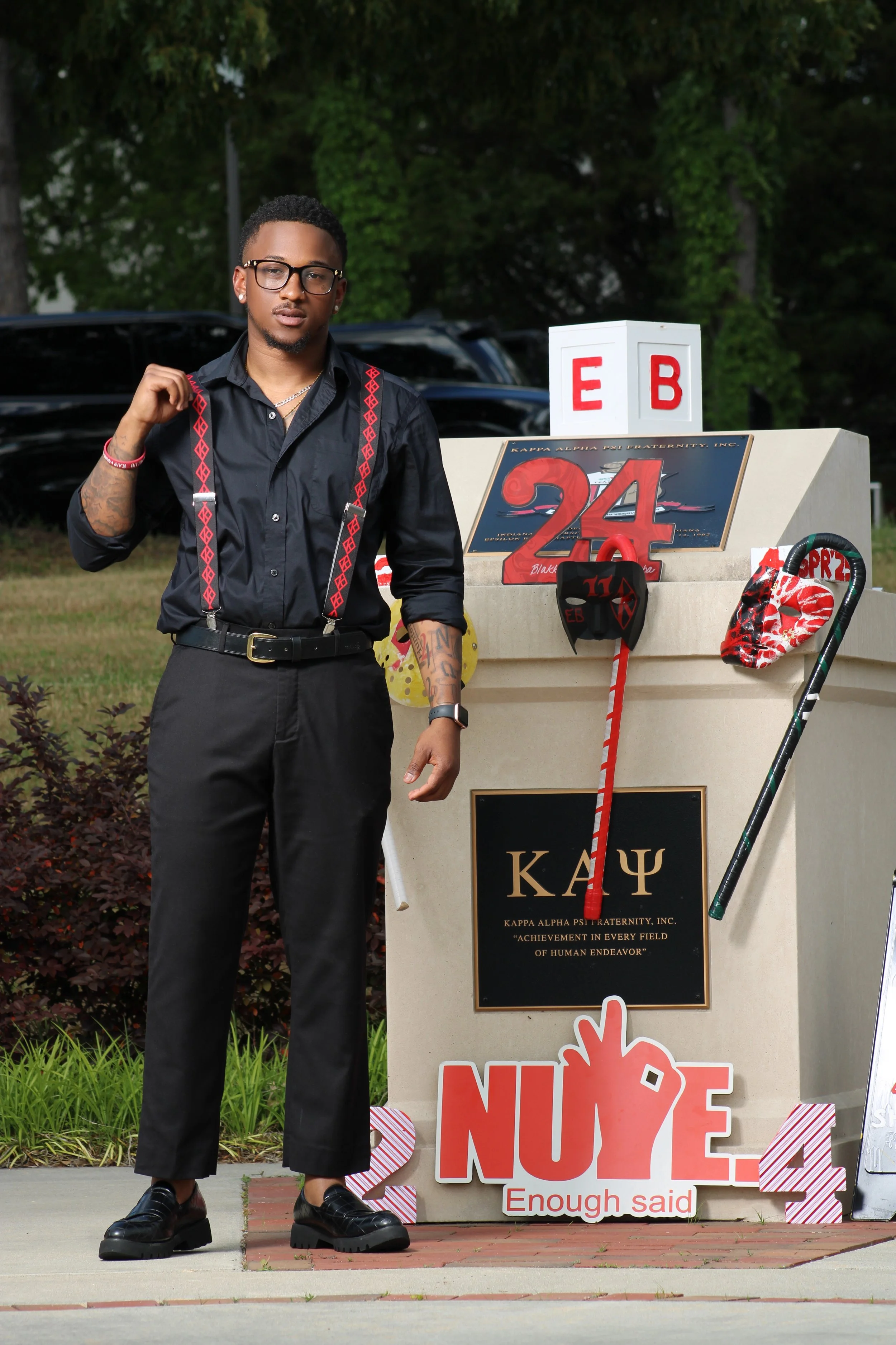 A young man standing outside near a fraternity sign with Greek letters, wearing glasses, a black shirt with red suspenders, and black pants. The sign features decorations related to a fraternity celebration.