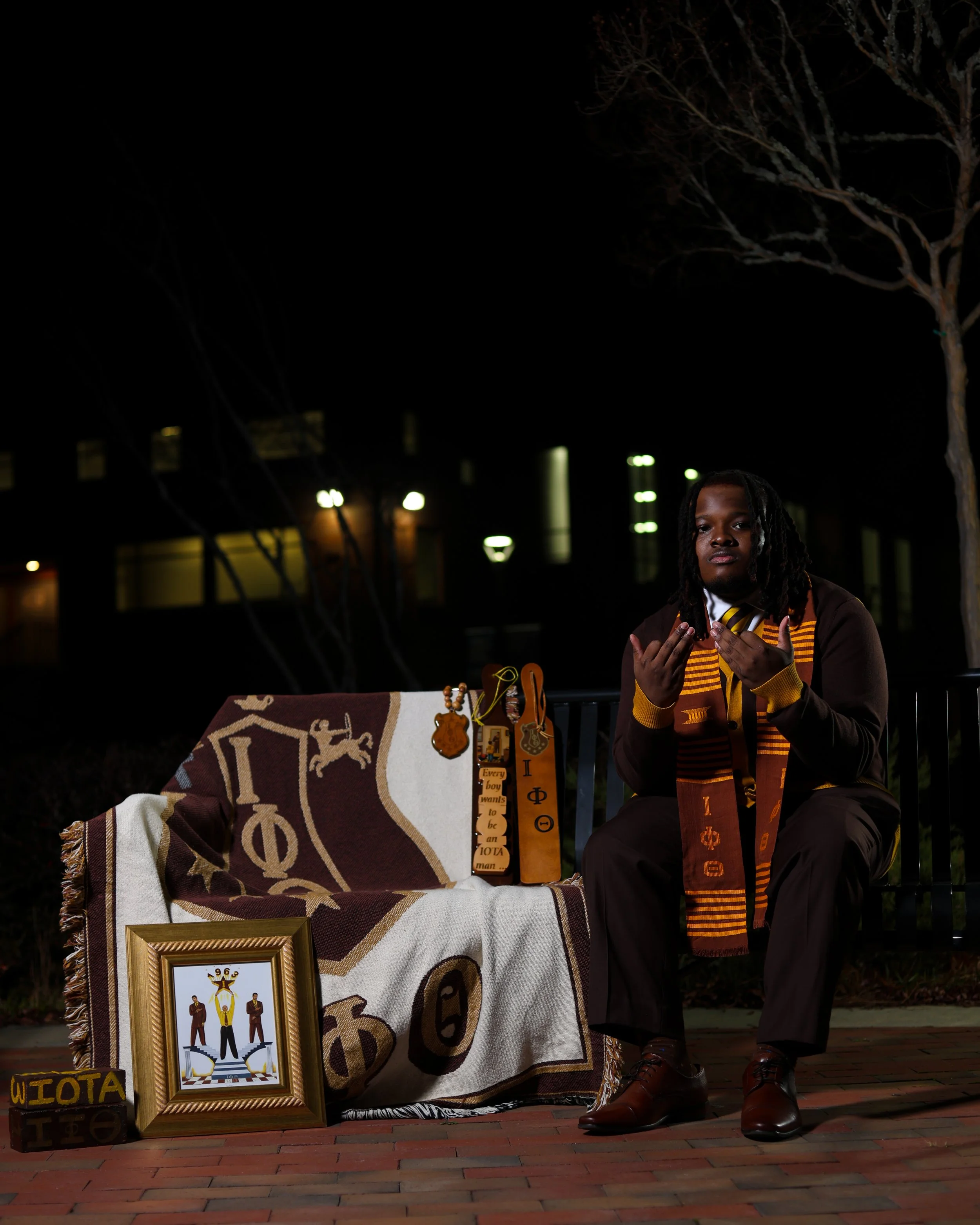 A person sitting on a bench displaying Greek life memorabilia, including a framed picture of a fraternity symbol, a blanket with Greek letters, and various keys and ribbons with Greek symbols. The person is wearing a fraternity sash and is outside at