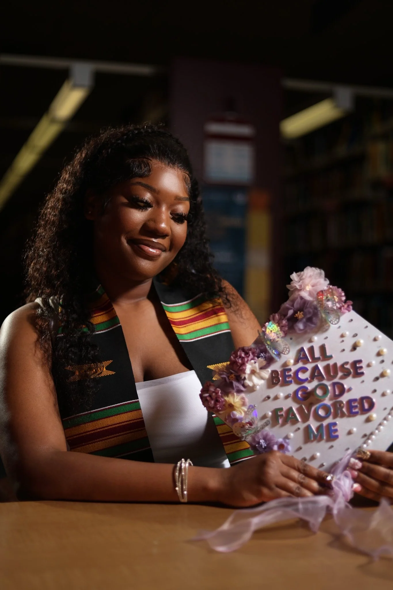 A woman in a white top and a colorful graduation sash sitting at a table, smiling while holding a decorated cake with the words 'All Because God Favored Me' written on it.