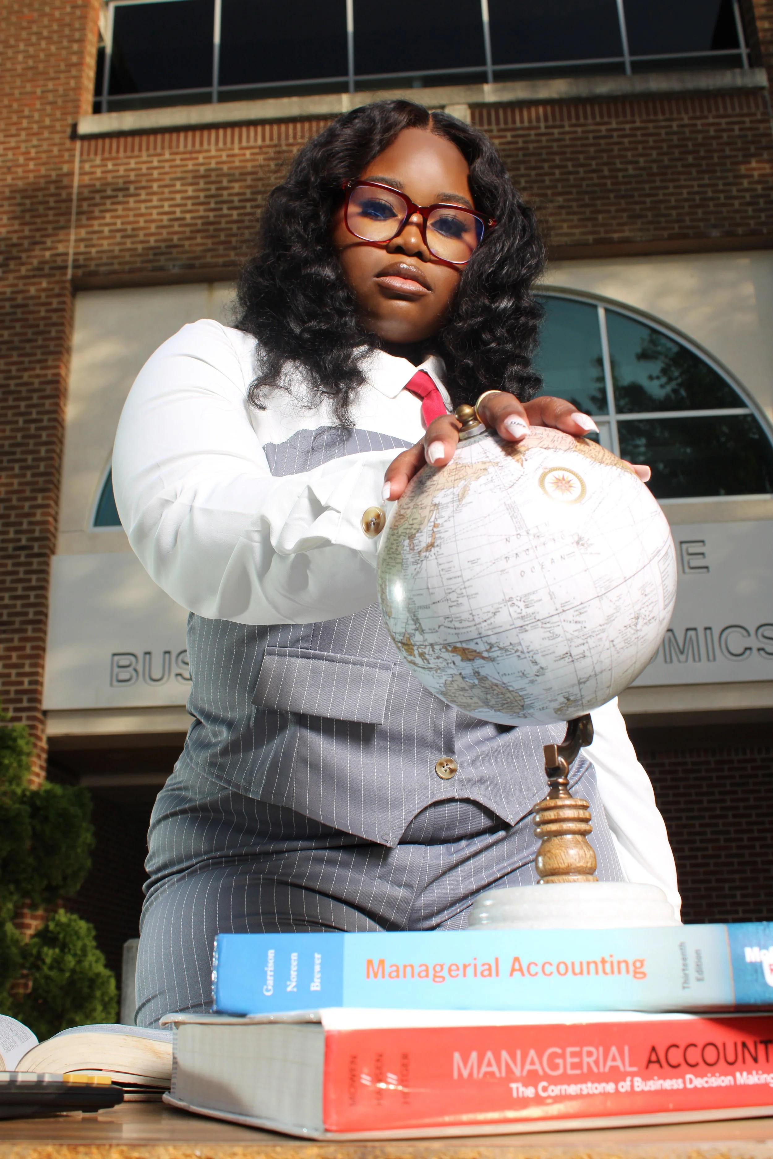 A woman in business attire holding a globe outdoors in front of a brick building with large windows.