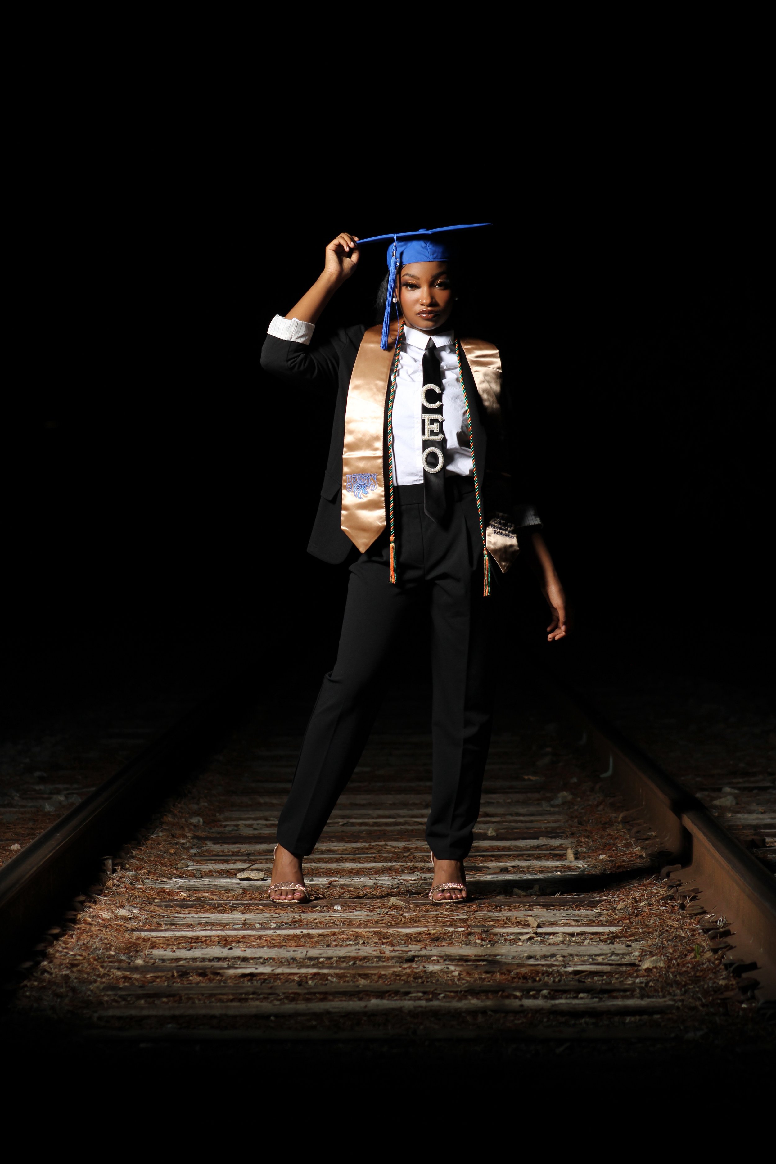 A woman in a graduation gown and cap standing on train tracks at night, holding her cap and wearing a sash and honor cords.