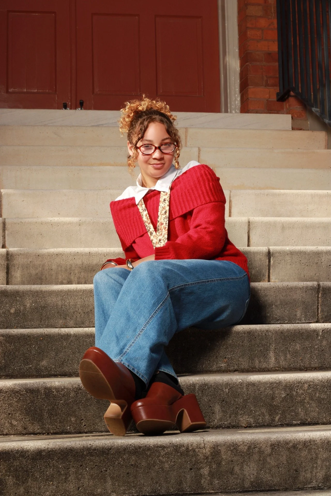 A young woman sitting on concrete stairs outside, wearing a red sweater, blue jeans, brown platform shoes, and glasses, with curly hair.