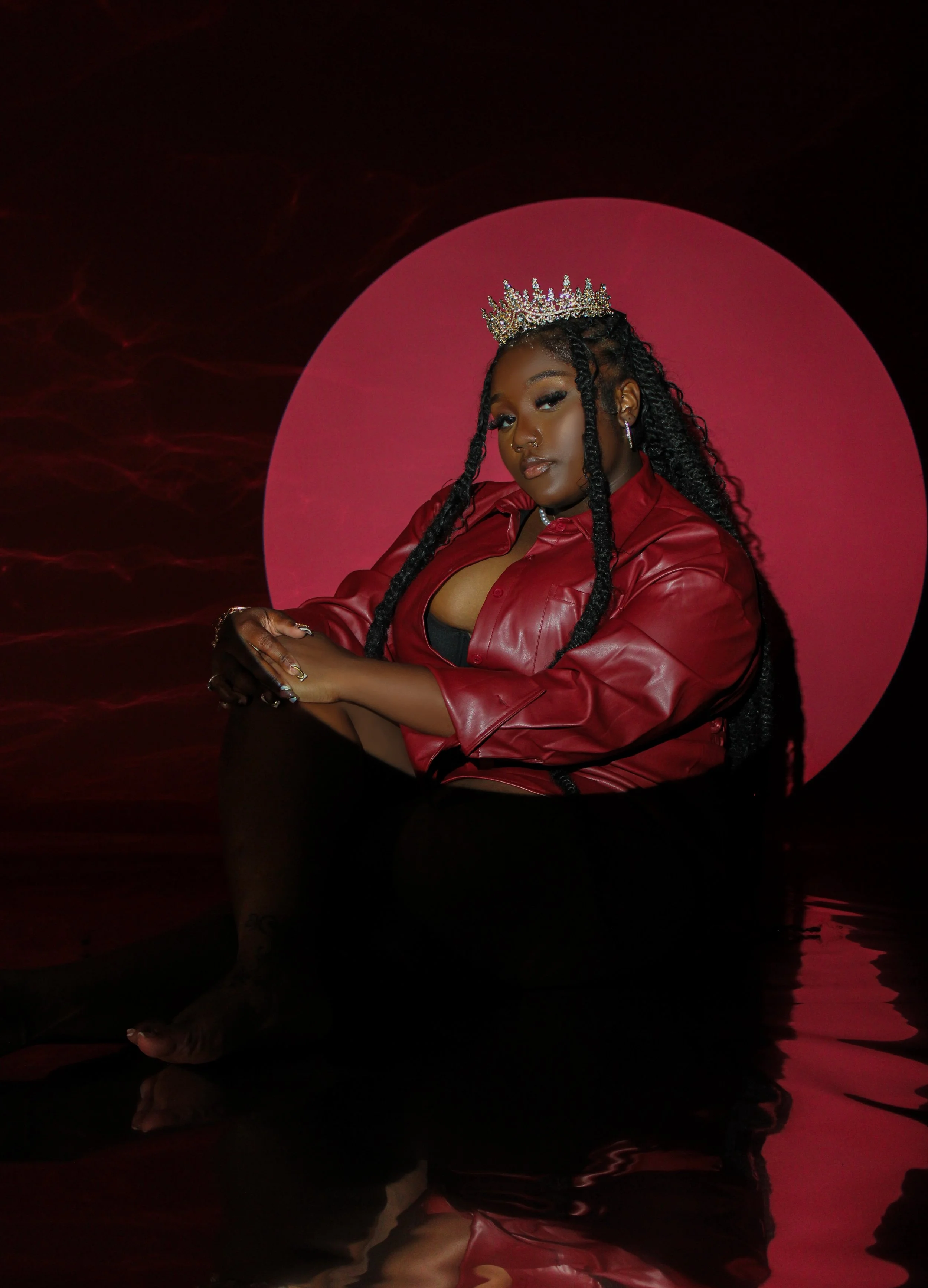 A woman with long twisted hair, wearing a red leather jacket and a crown, sitting with her legs crossed in front of a pink circle background. She has bold makeup and jewelry.