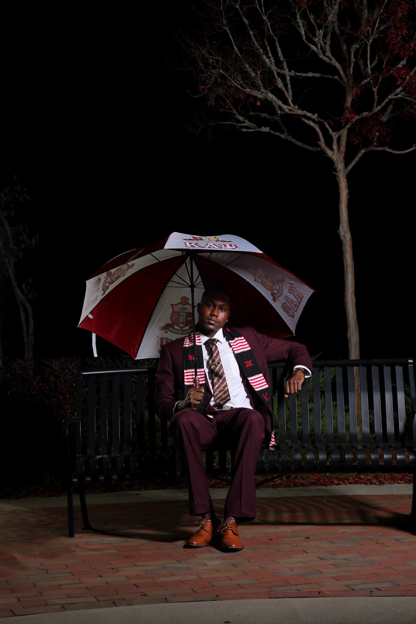 A man in a maroon suit sits on a black park bench at night, holding an umbrella with sports team logos, wearing a tie and a scarf, with leafless trees and a dark sky in the background.