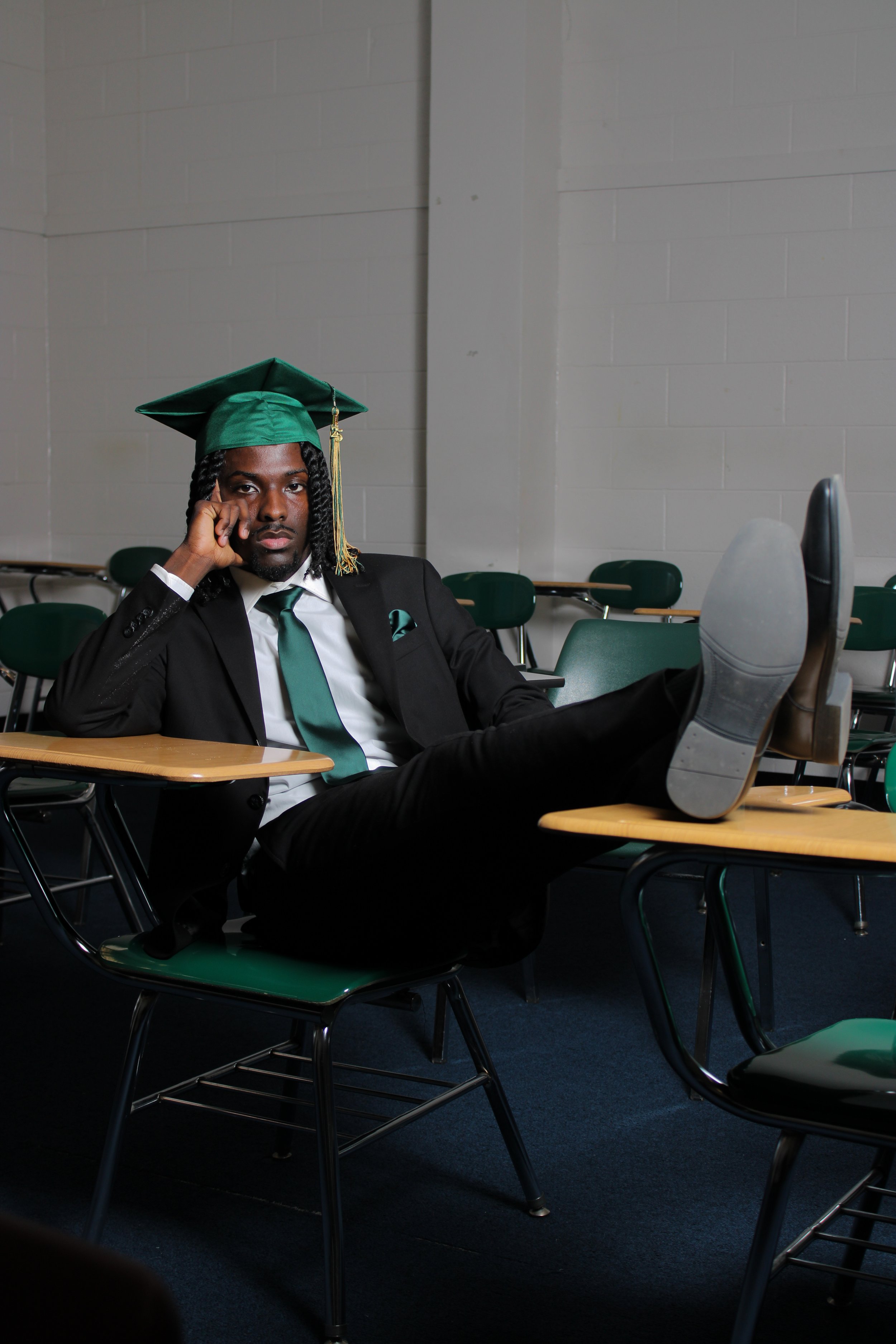 A man in a black suit, wearing a green graduation cap and gown, sitting in a classroom with empty desks, with his legs crossed on a desk, looking at the camera with his hand resting on his face.