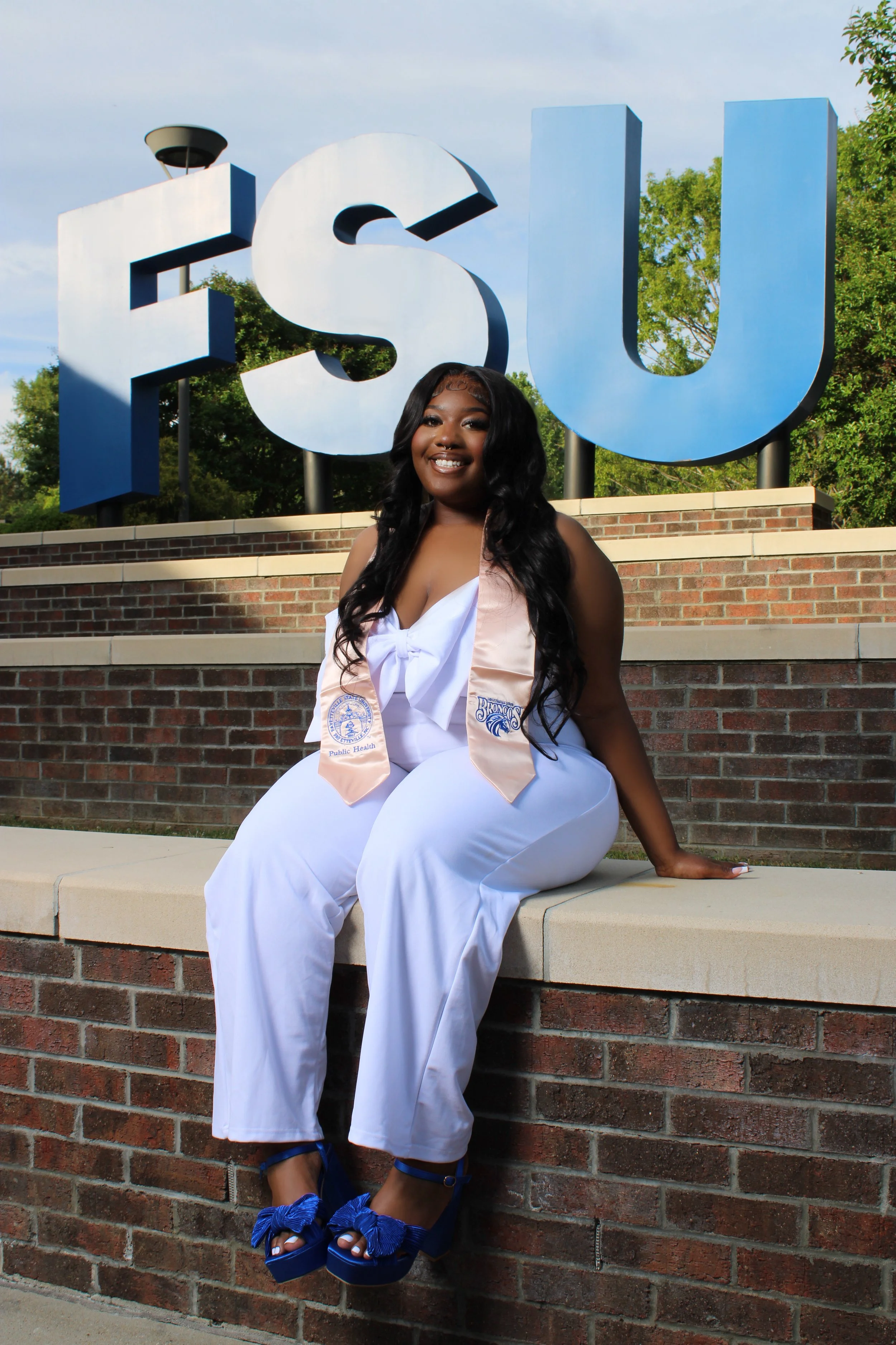 A young woman in a white graduation dress and blue high heels, sitting on a brick wall in front of a large 'FSU' sign, smiling at the camera during her graduation celebration.