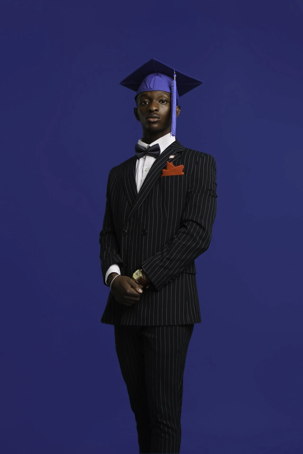 Young man in a black pinstripe suit, white shirt, black bow tie, red pocket square, wearing a blue graduation cap with tassel, standing against a solid blue background.
