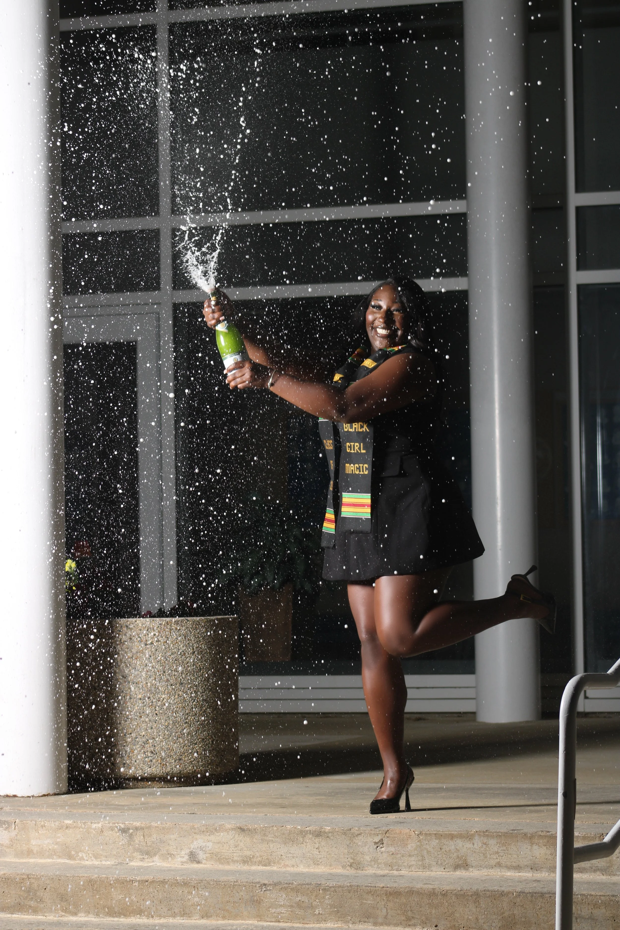 Woman celebrating with a champagne bottle, spraying it open, with champagne foam in the air, smiling, wearing a black dress, high heels, and a sash that reads 'Black Girl Magic', standing outdoors near a building.