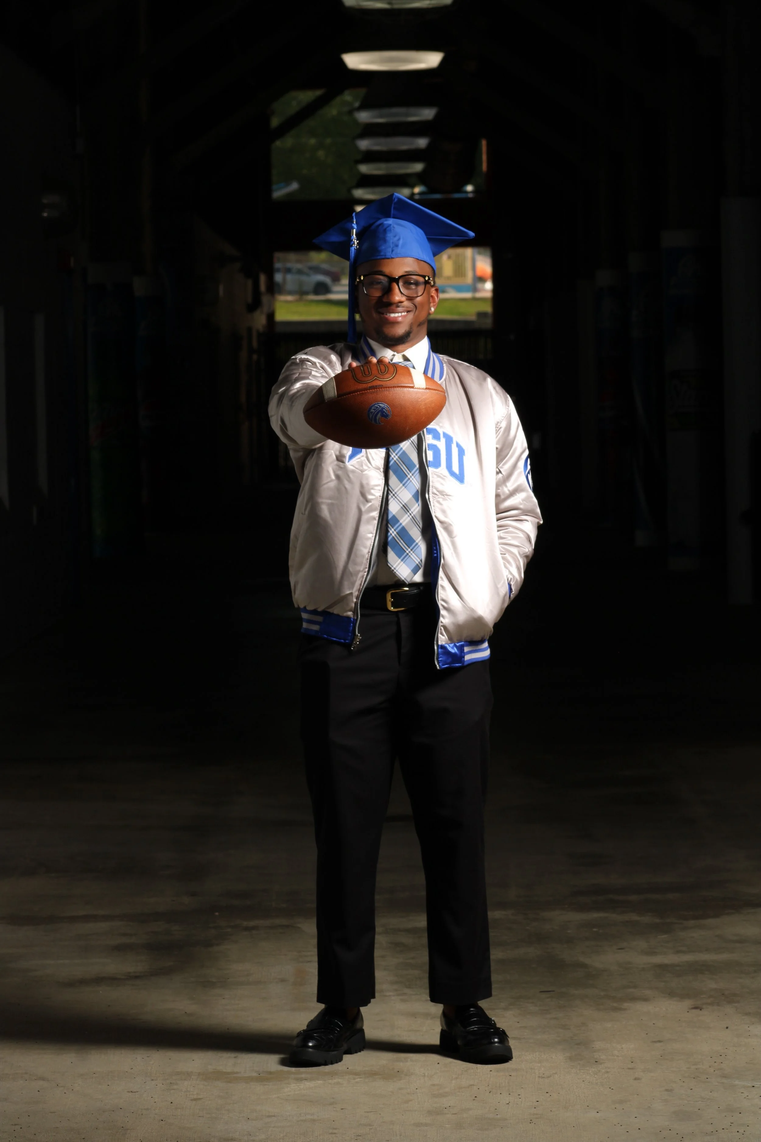 Young man in graduation cap and gown holding football, standing in dimly lit tunnel, smiling.