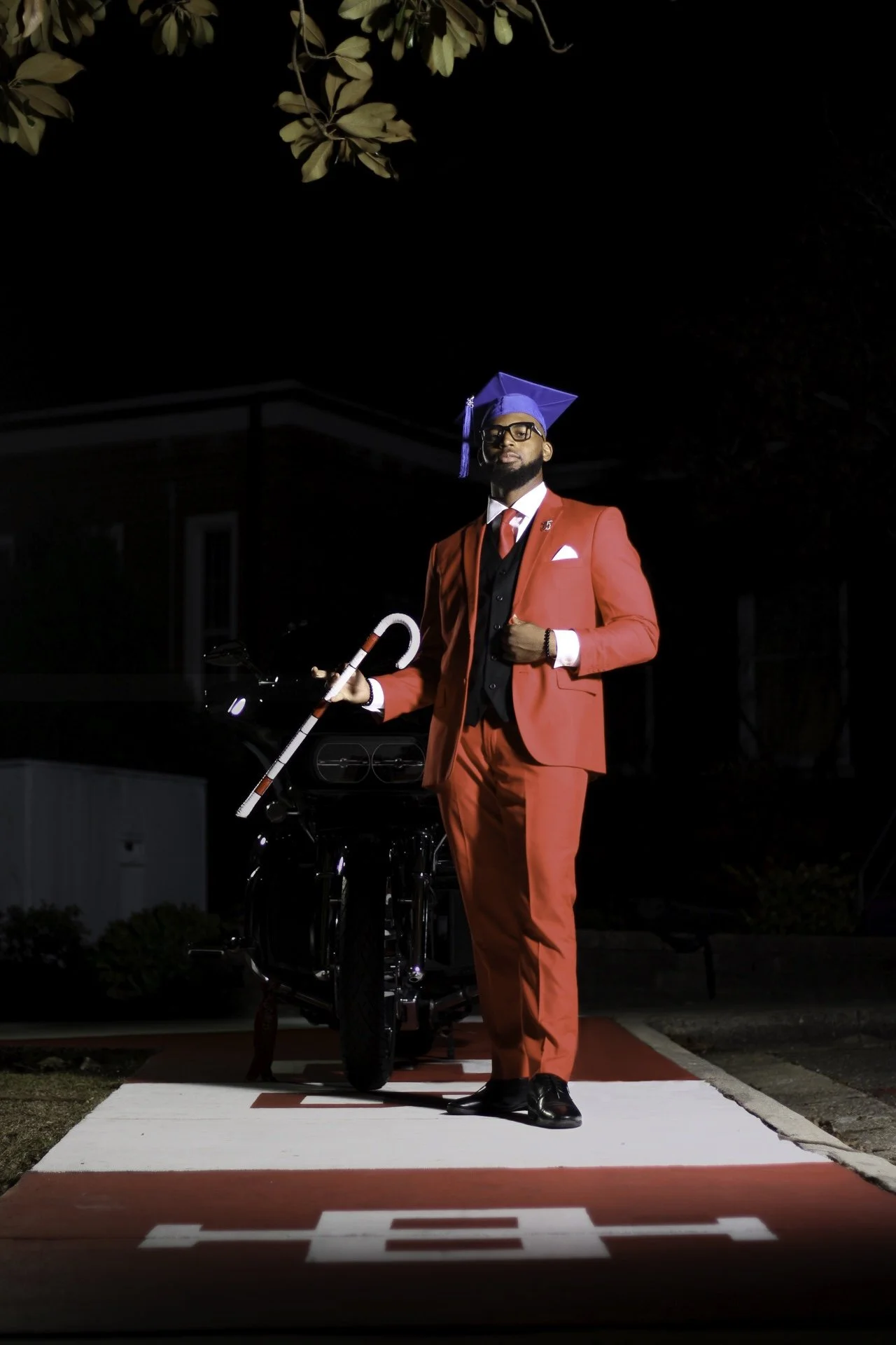 A man dressed in a red suit and black shoes, wearing a blue graduation cap, standing on a red and white mat outdoors at night. He is holding a white cane with a red stripe and standing next to a black motorcycle. The background is dark with some foli