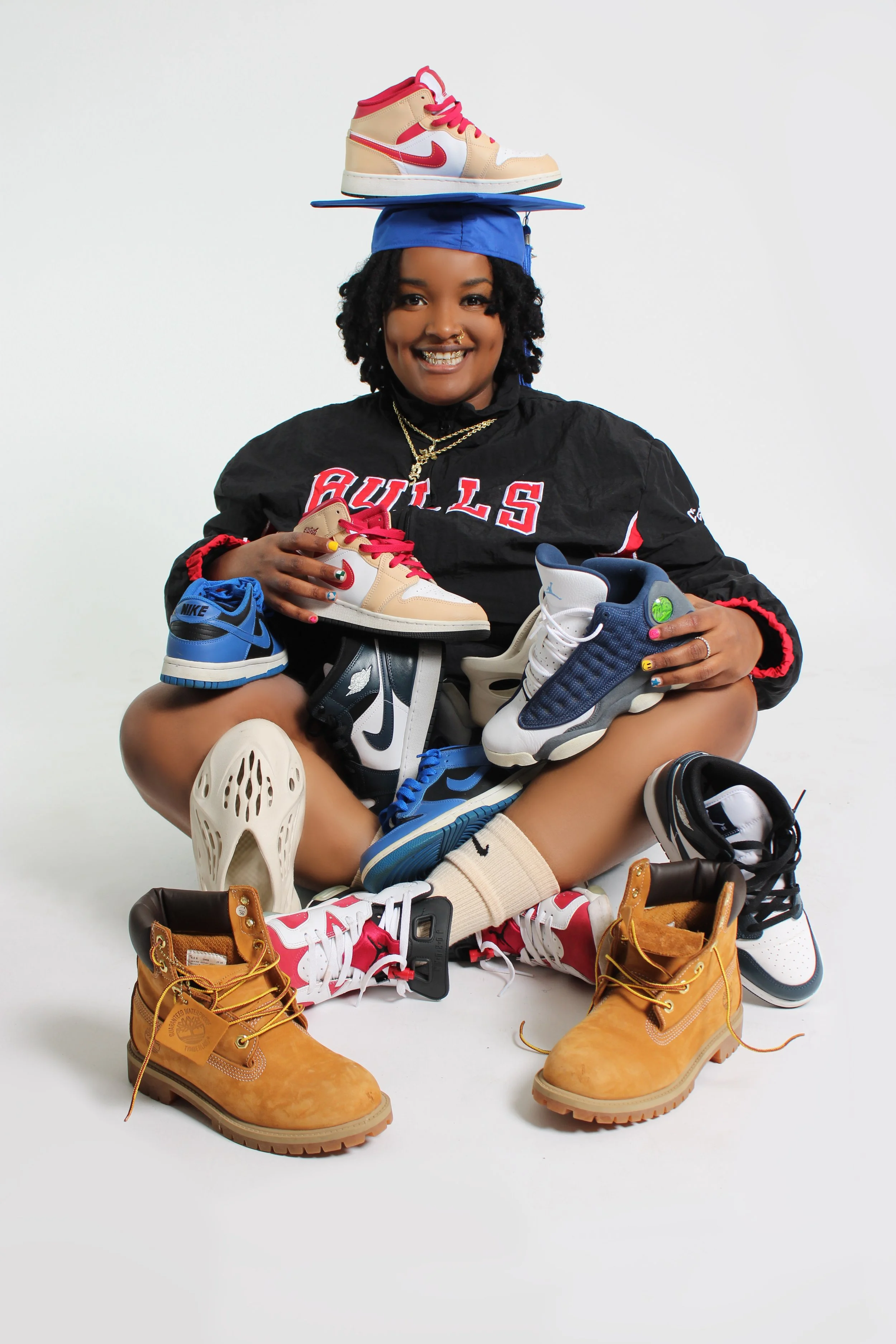 Person sitting surrounded by various sneakers, wearing a blue graduation cap, a black jersey, and red and white sneakers.