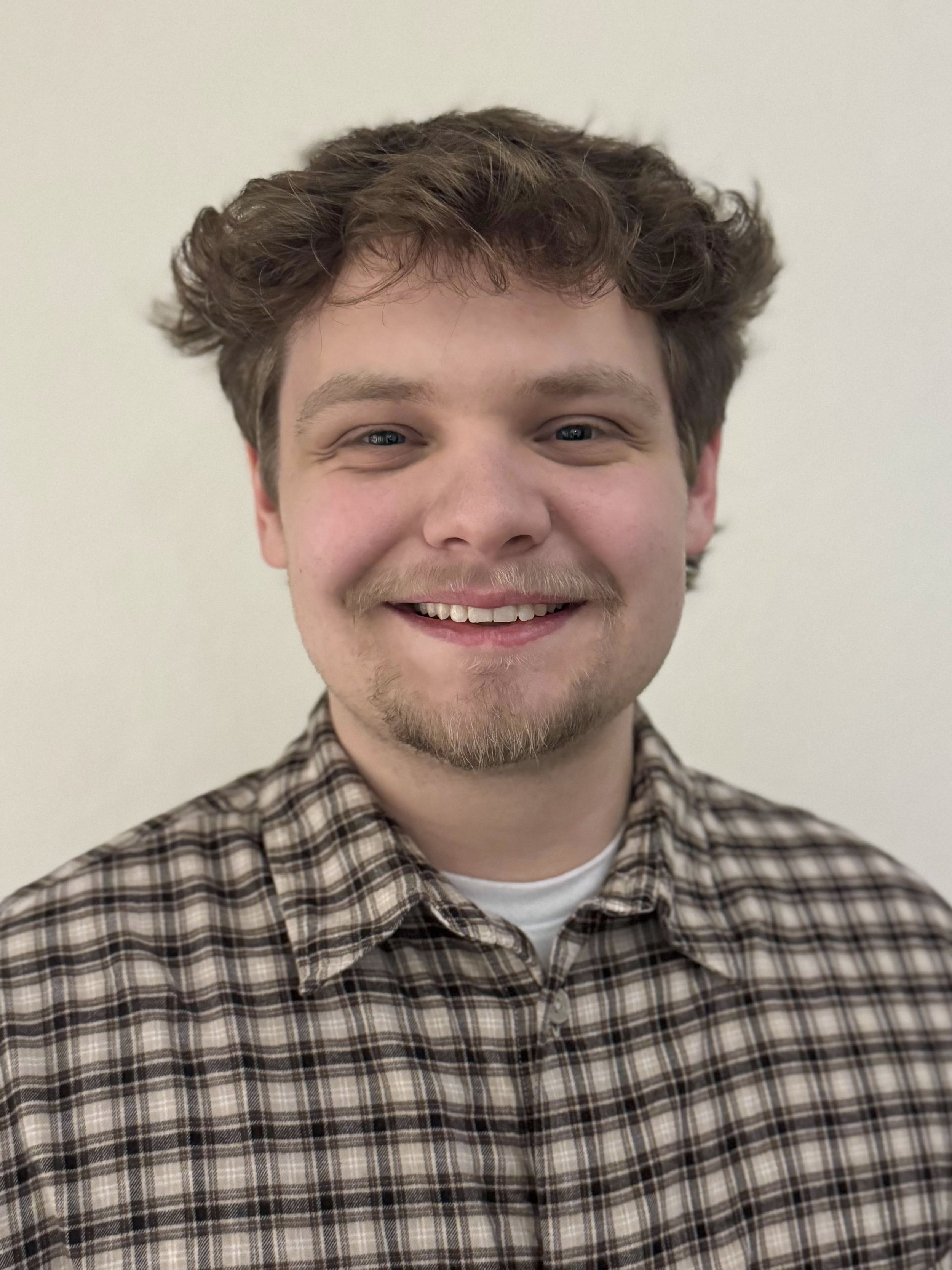 A young man with wavy brown hair, blue eyes, and a light beard, smiling at the camera against a plain white wall backdrop, wearing a brown plaid shirt.