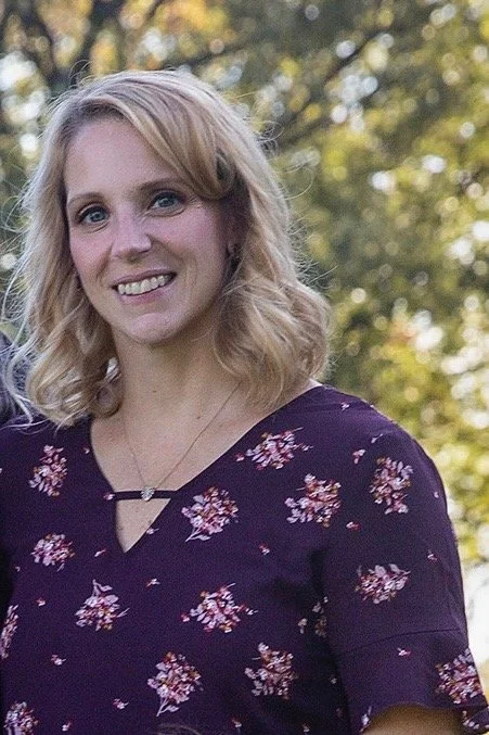 A smiling woman with shoulder-length blonde hair posing outdoors, wearing a dark purple floral dress.