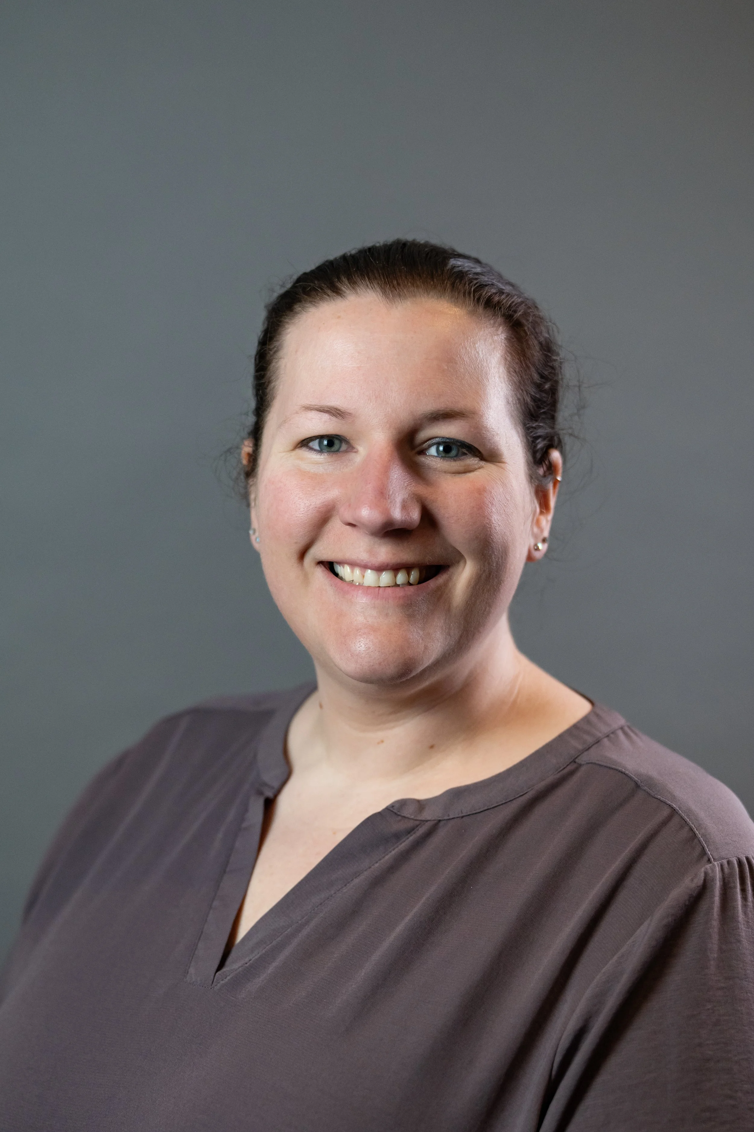 A woman with brown hair tied back, smiling, wearing a brown top with a V-neck, against a gray background.